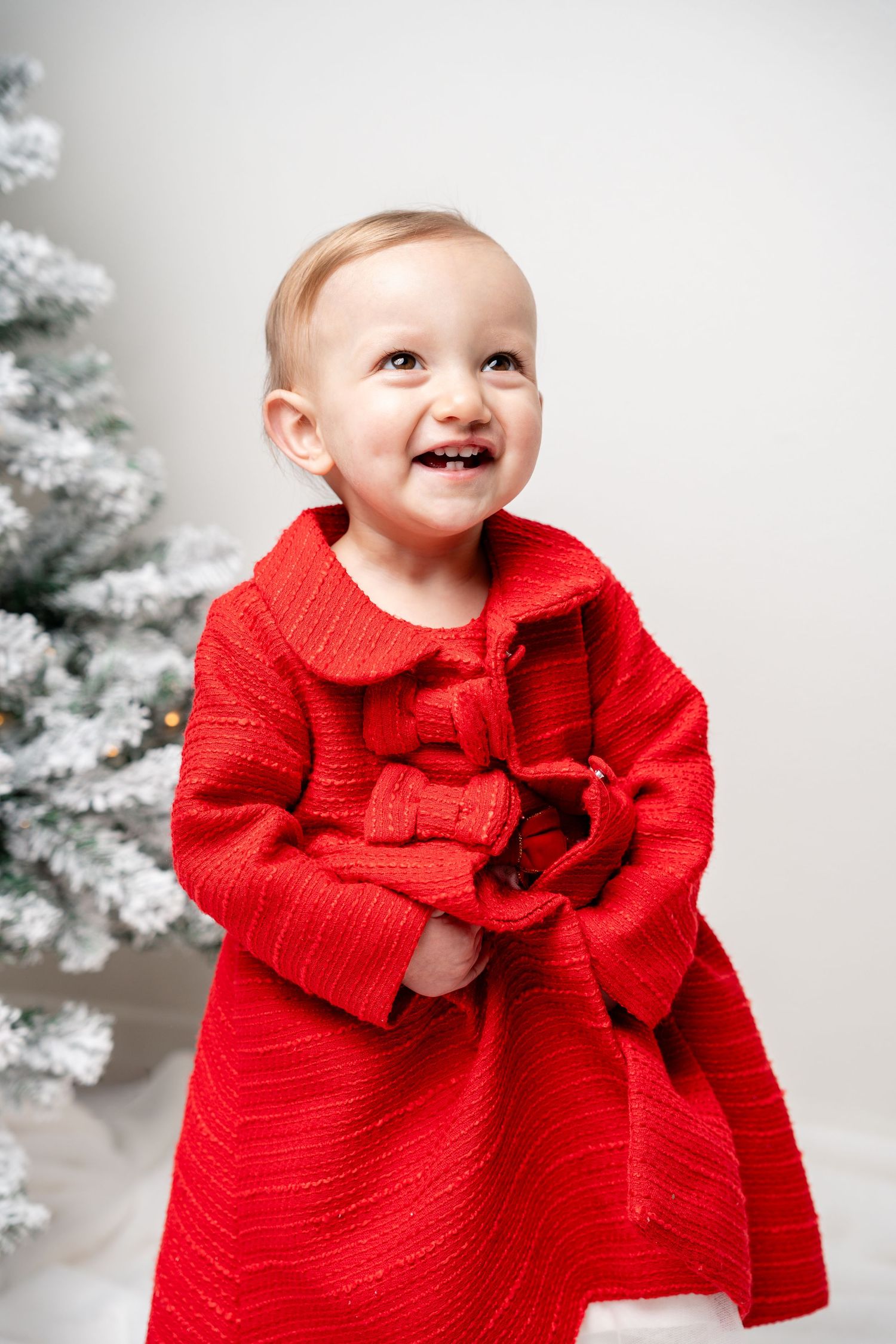 Smiling baby girl in red coat standing near a snow-flocked Christmas tree.