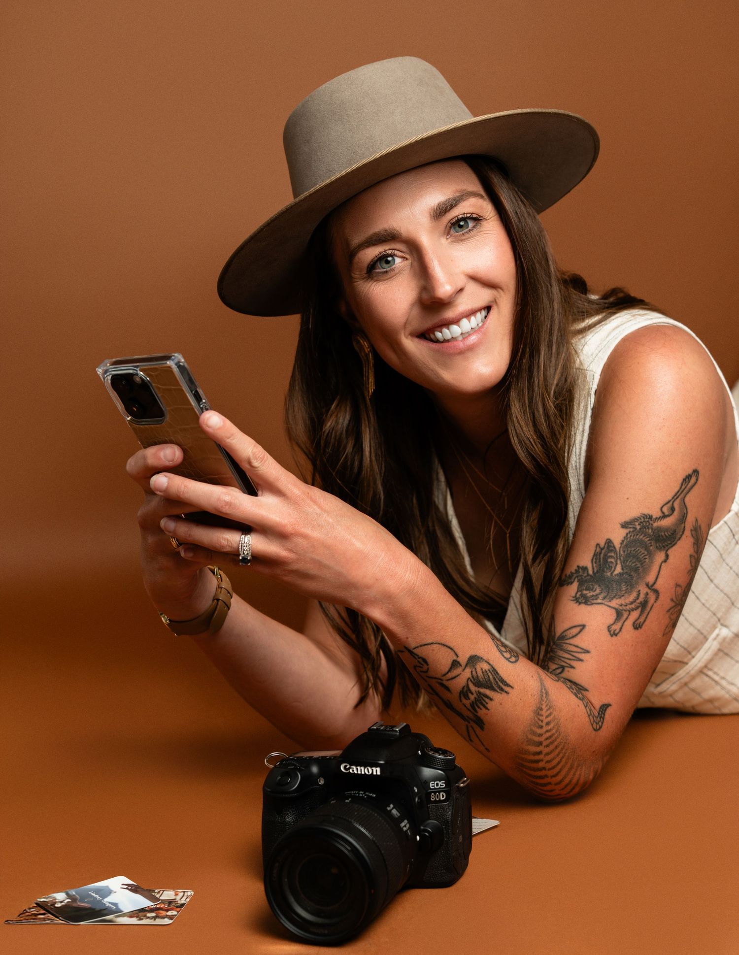Photographer with tattoos wearing a tan hat poses with camera and phone on brown background.