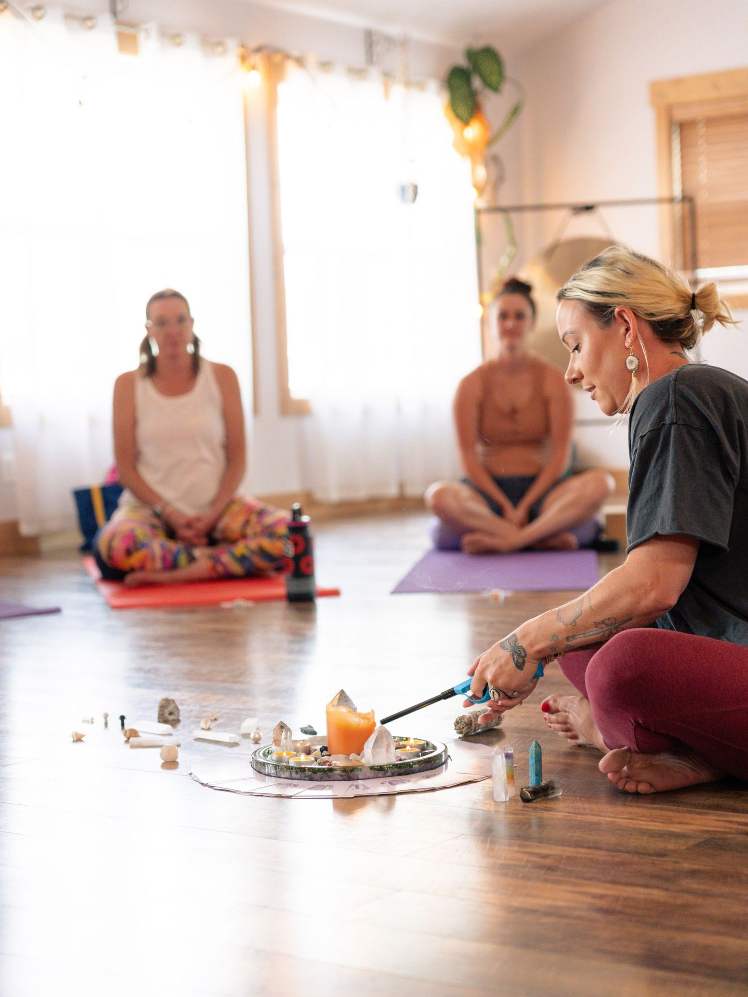 Meditation class participants sit on yoga mats while instructor arranges spiritual items on wooden floor.