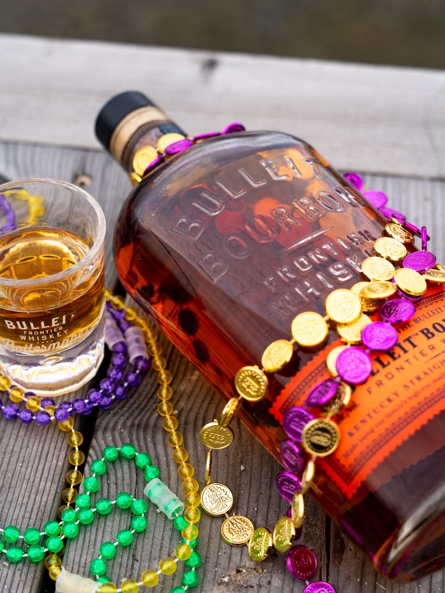 Bottle of Bulleit bourbon decorated with colorful Mardi Gras beads on wooden surface with whiskey glass.