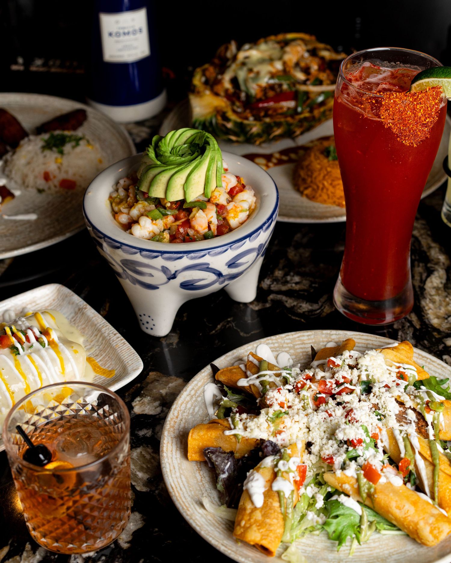 Festive spread of Mexican dishes with colorful cocktails and garnished taquitos on dark table setting.