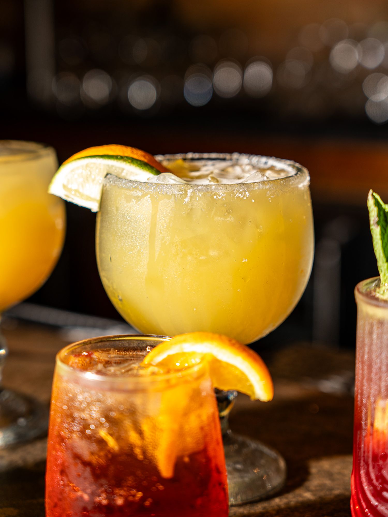 Colorful cocktails garnished with orange slices featuring yellow and red drinks in margarita glasses.