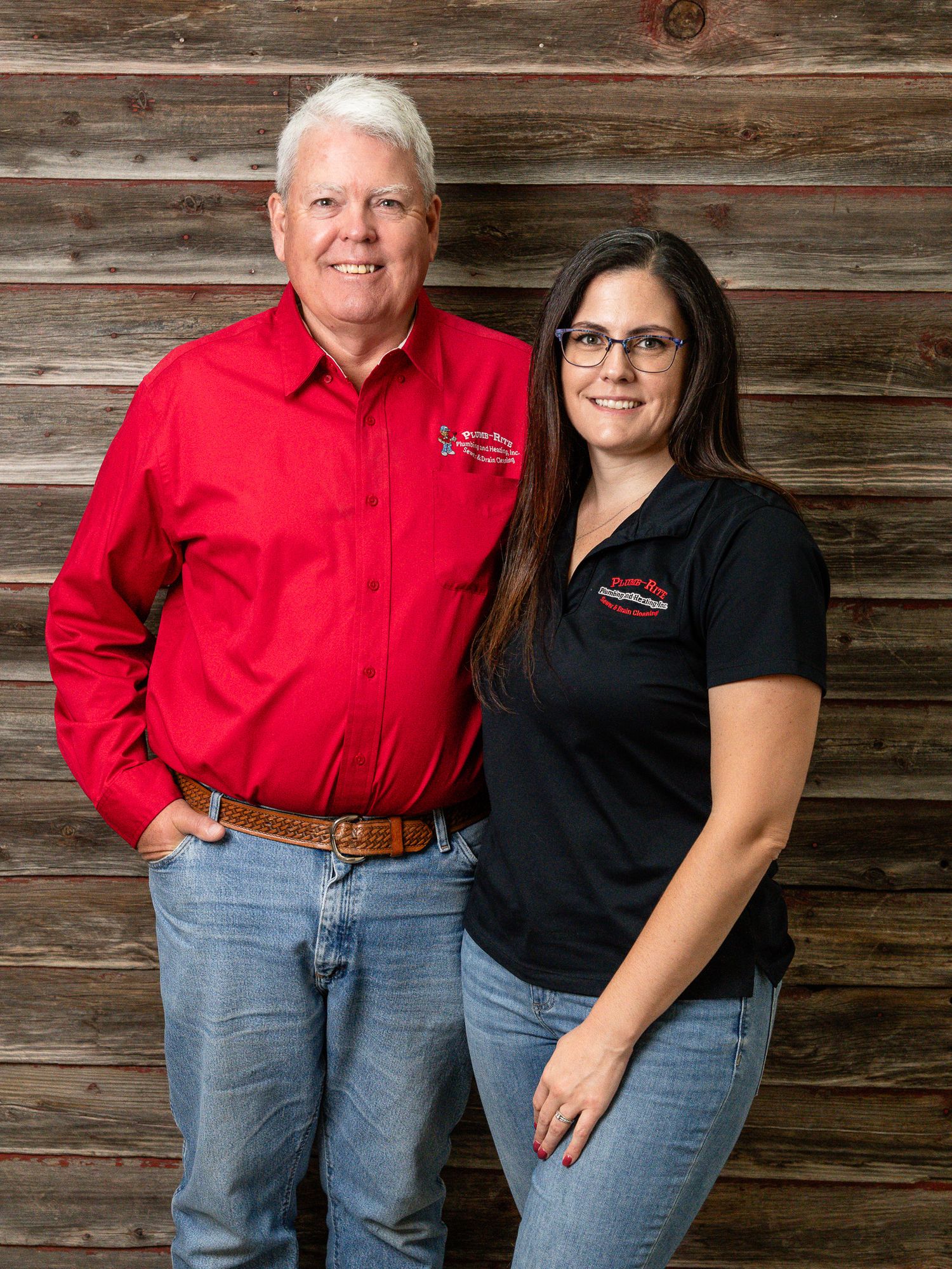 Two coworkers in branded company uniforms pose against rustic wood wall.