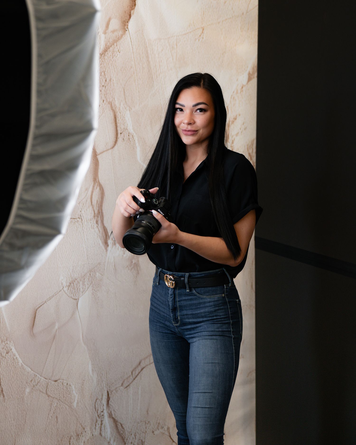 Photographer in black shirt and jeans holding camera against textured wall.