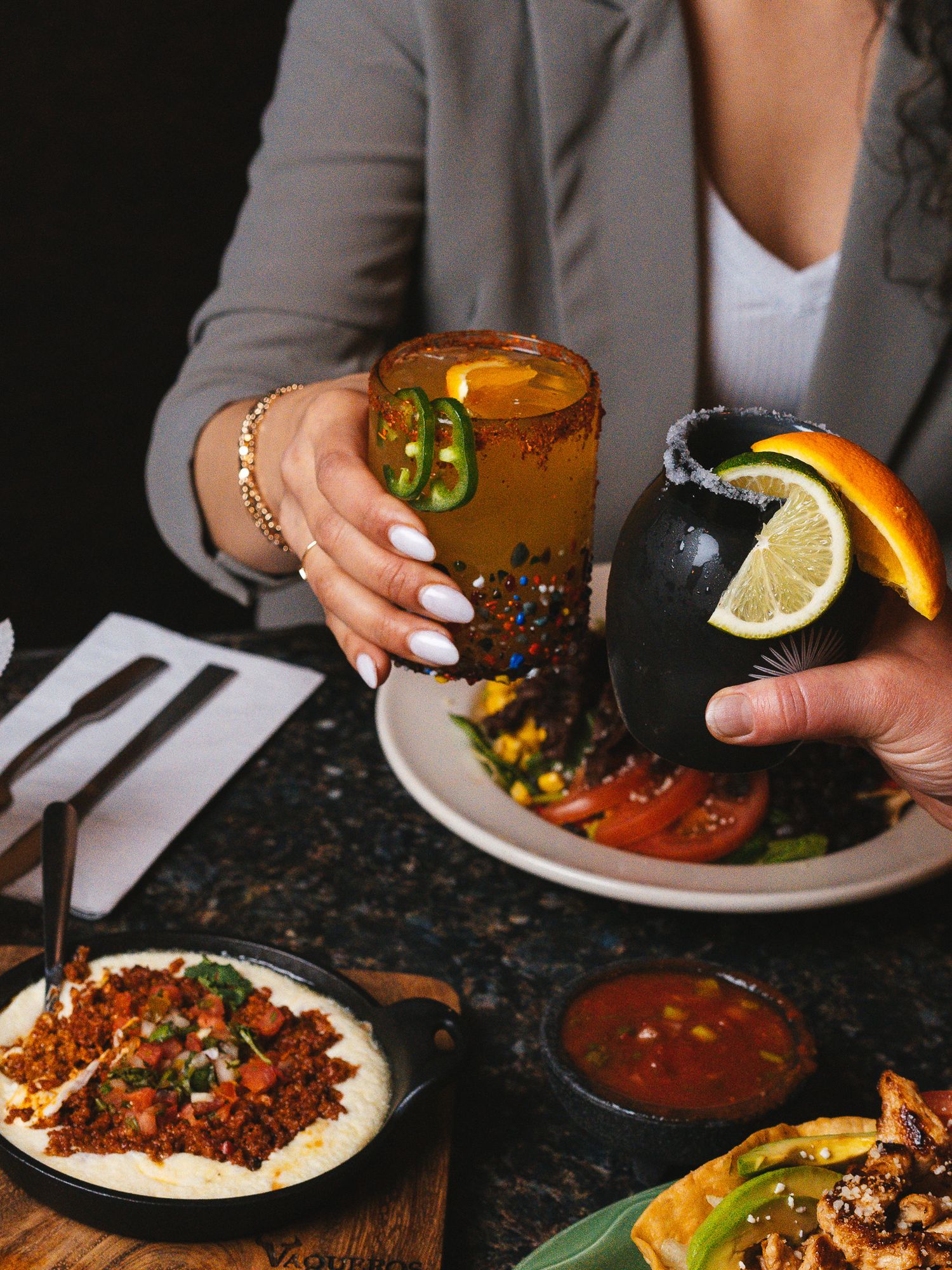 Hands holding spicy cocktails garnished with jalapenos and orange slices alongside Mexican dishes.