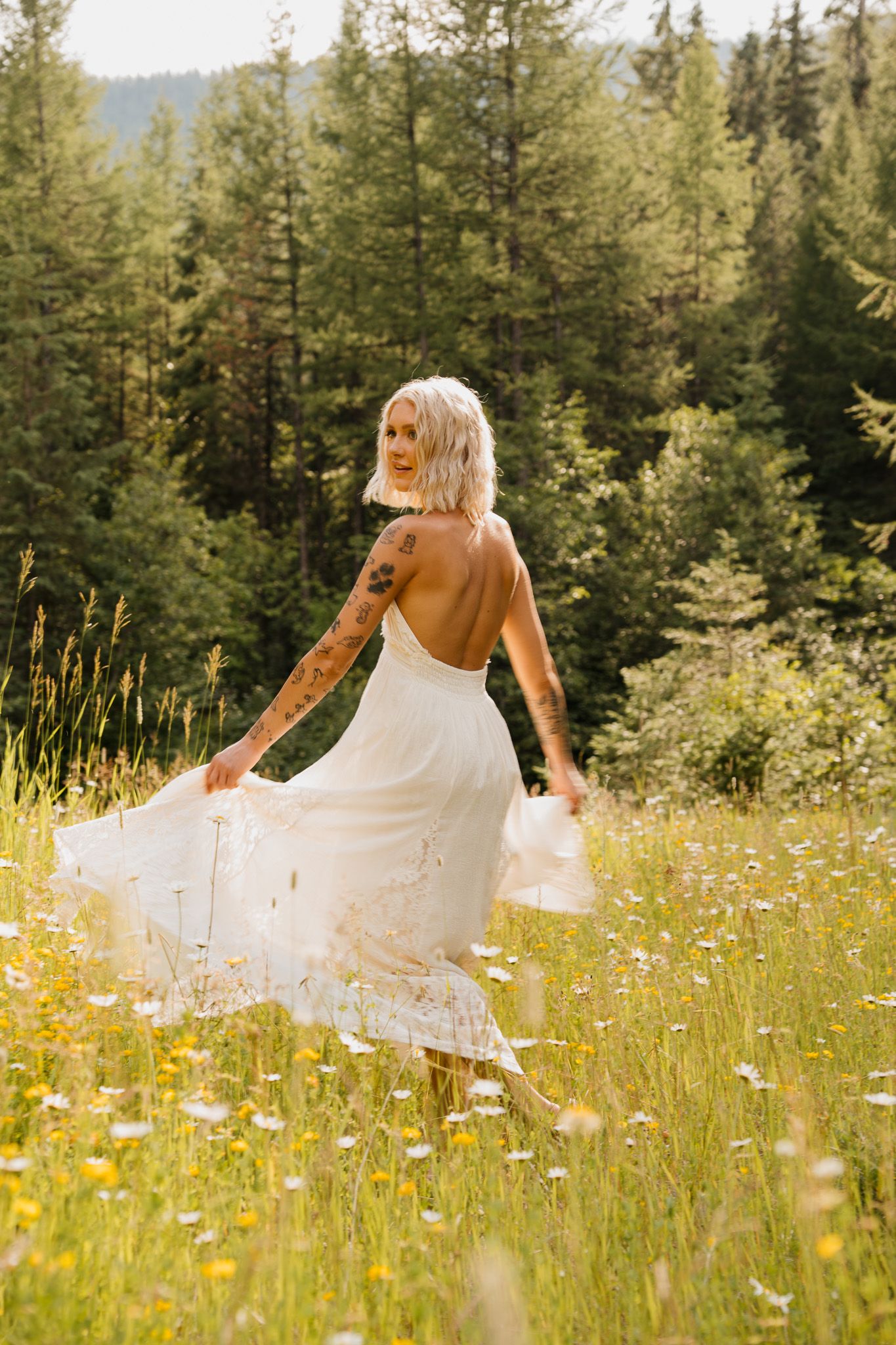 Tattooed bride in backless white lace dress twirling in wildflower meadow with evergreen forest backdrop.