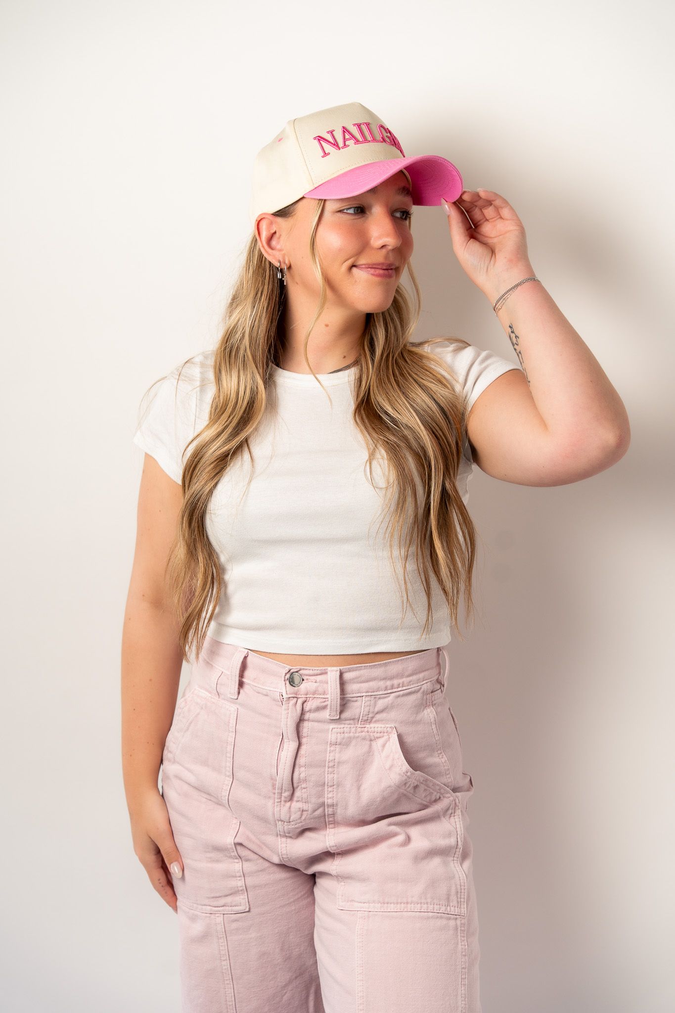 Woman in white t-shirt and pink pants wearing a pink baseball cap with 'NAILS' text.