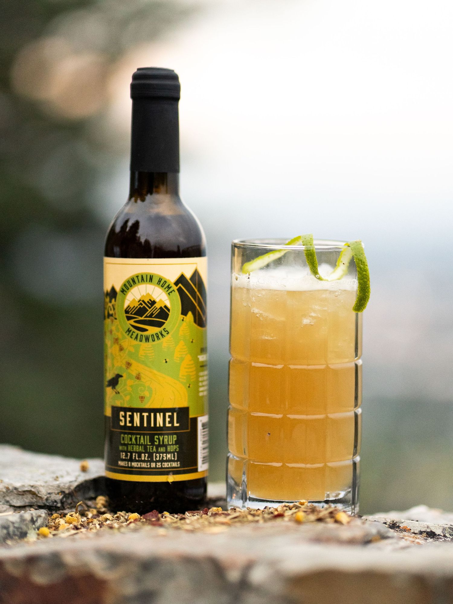 Bottle of Sunshine Elixir with matching citrus cocktail garnished with lime on rocky ledge.