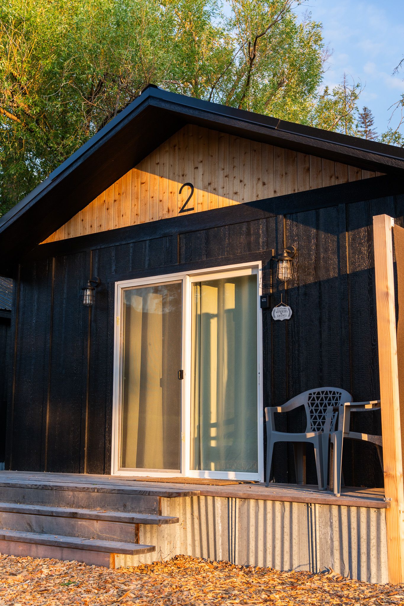 A small black cabin with wooden accent trim and number 2 features a sliding glass door and plastic chair on the porch.