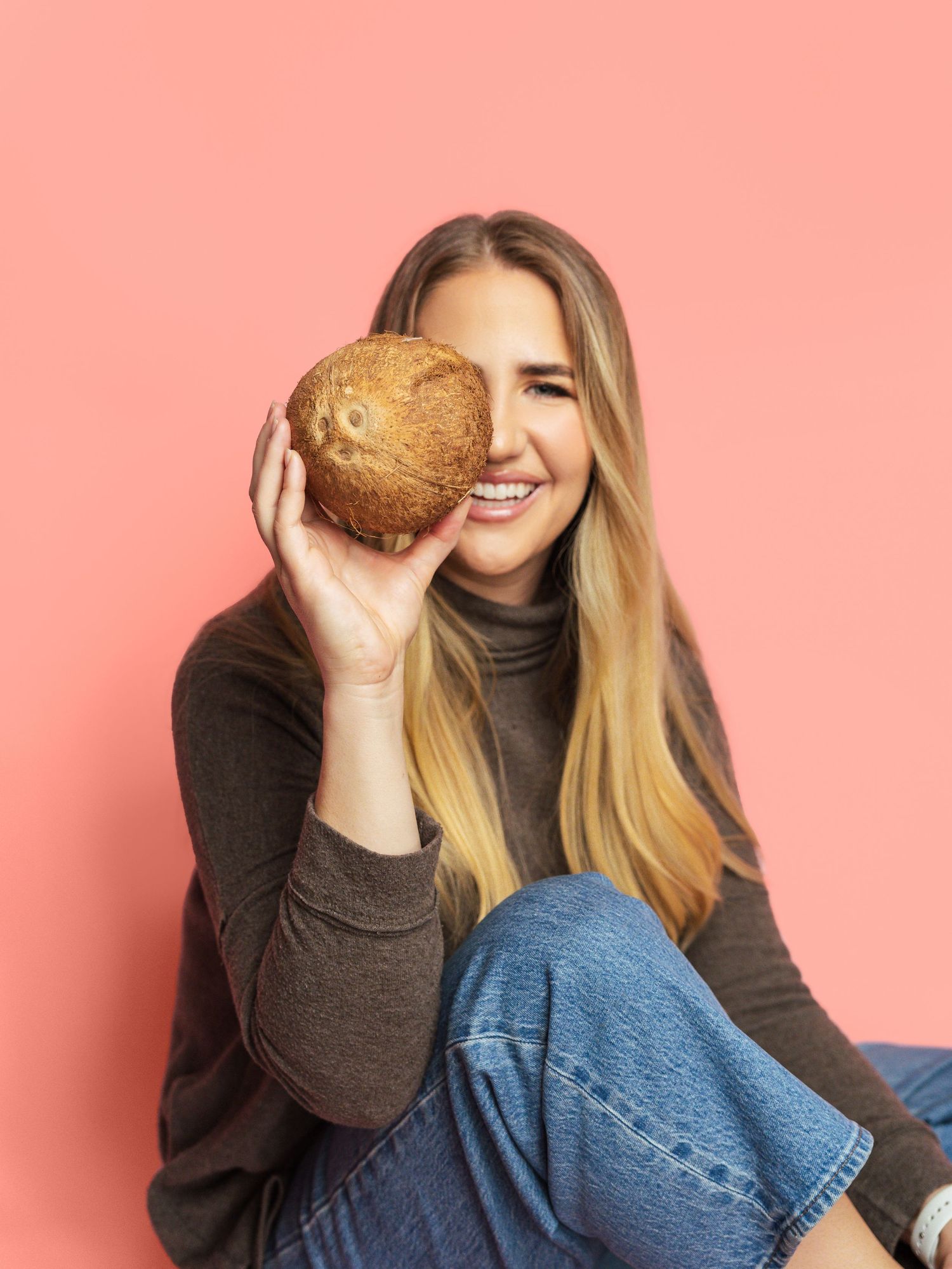 Person in black sweater holding coconut against coral pink backdrop.