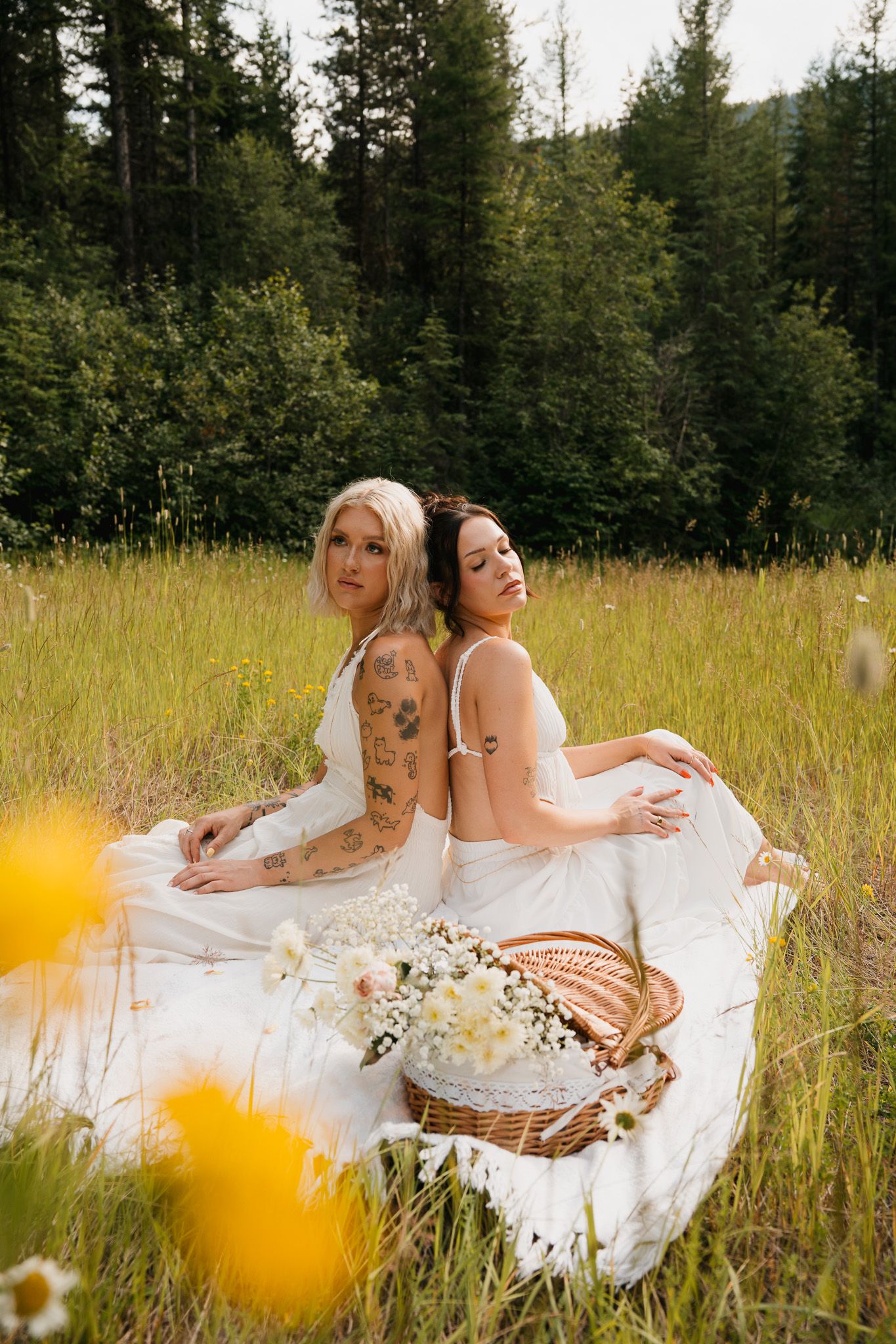 Two women in white dresses sit back-to-back in a meadow with wildflowers and a wicker basket, forest backdrop.