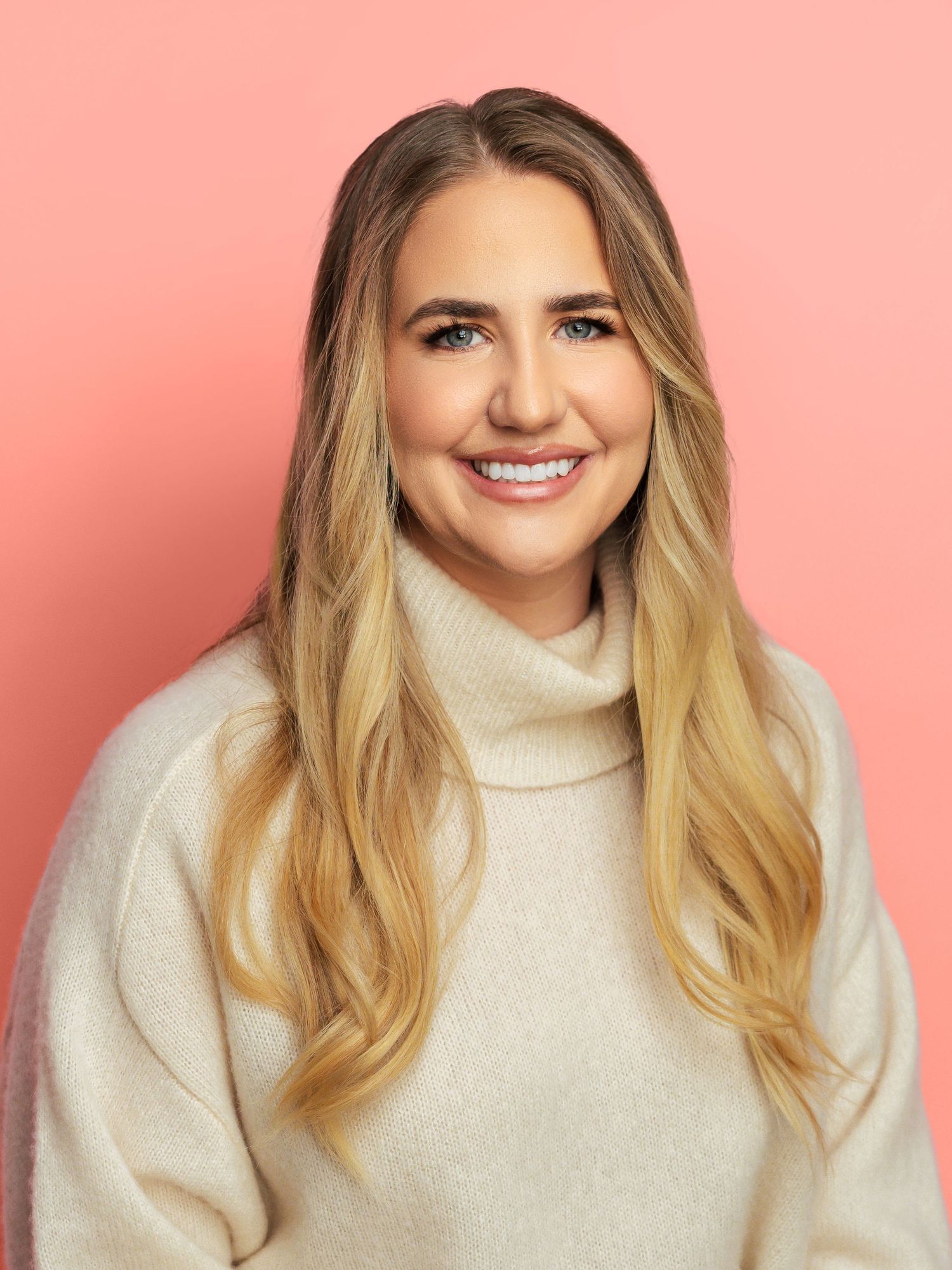 A person in a beige turtleneck sweater photographed against a pink background.