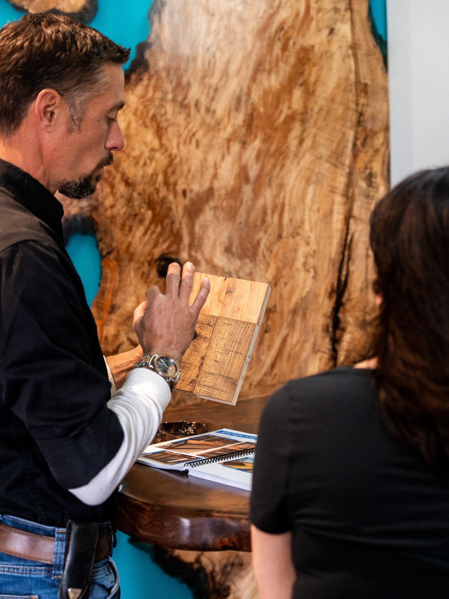 Two people examining wood samples and reference materials against a turquoise blue wall background.