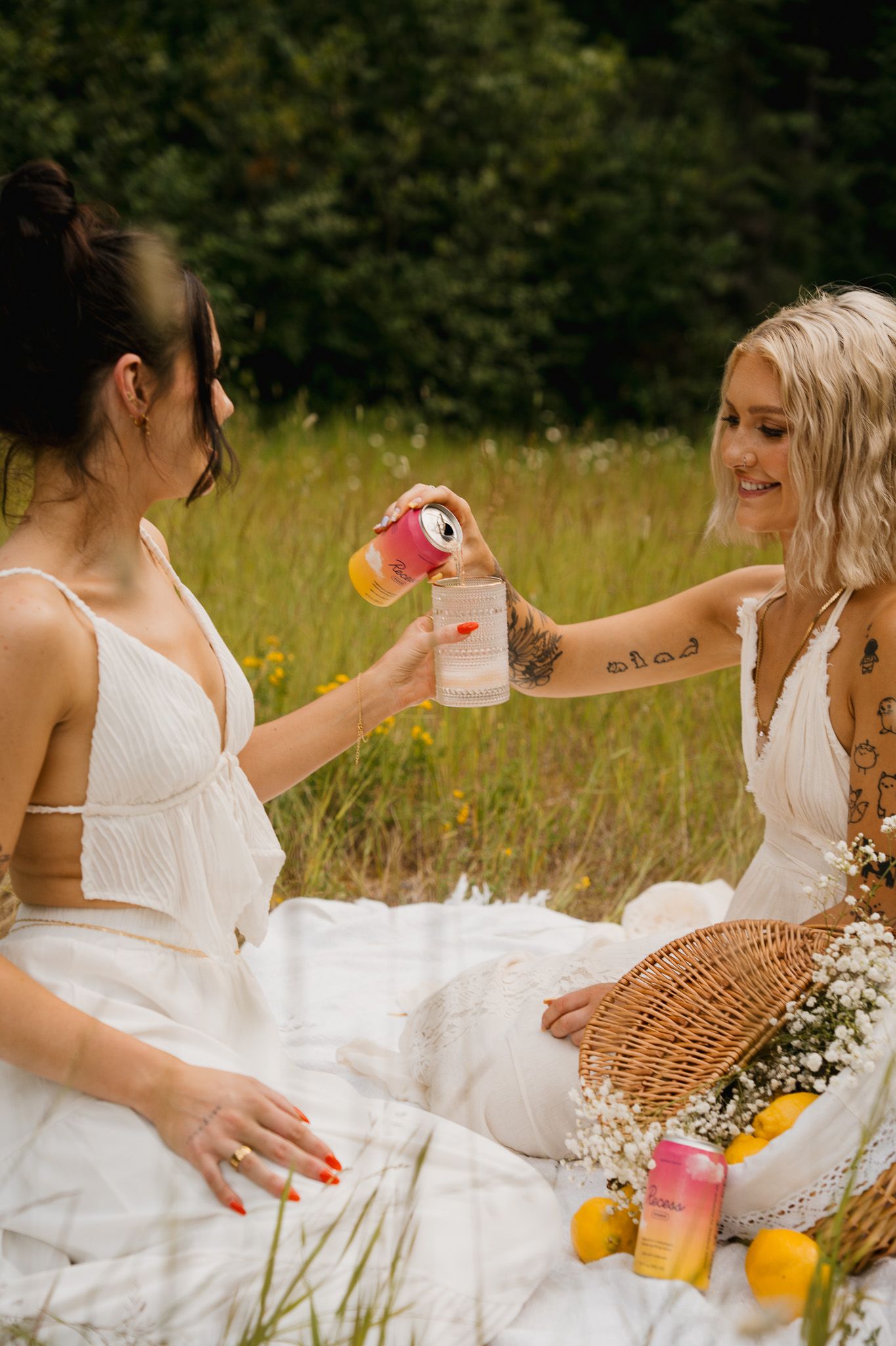 Women in white dresses enjoy a picnic on a sunny meadow with colorful drinks and wicker baskets.