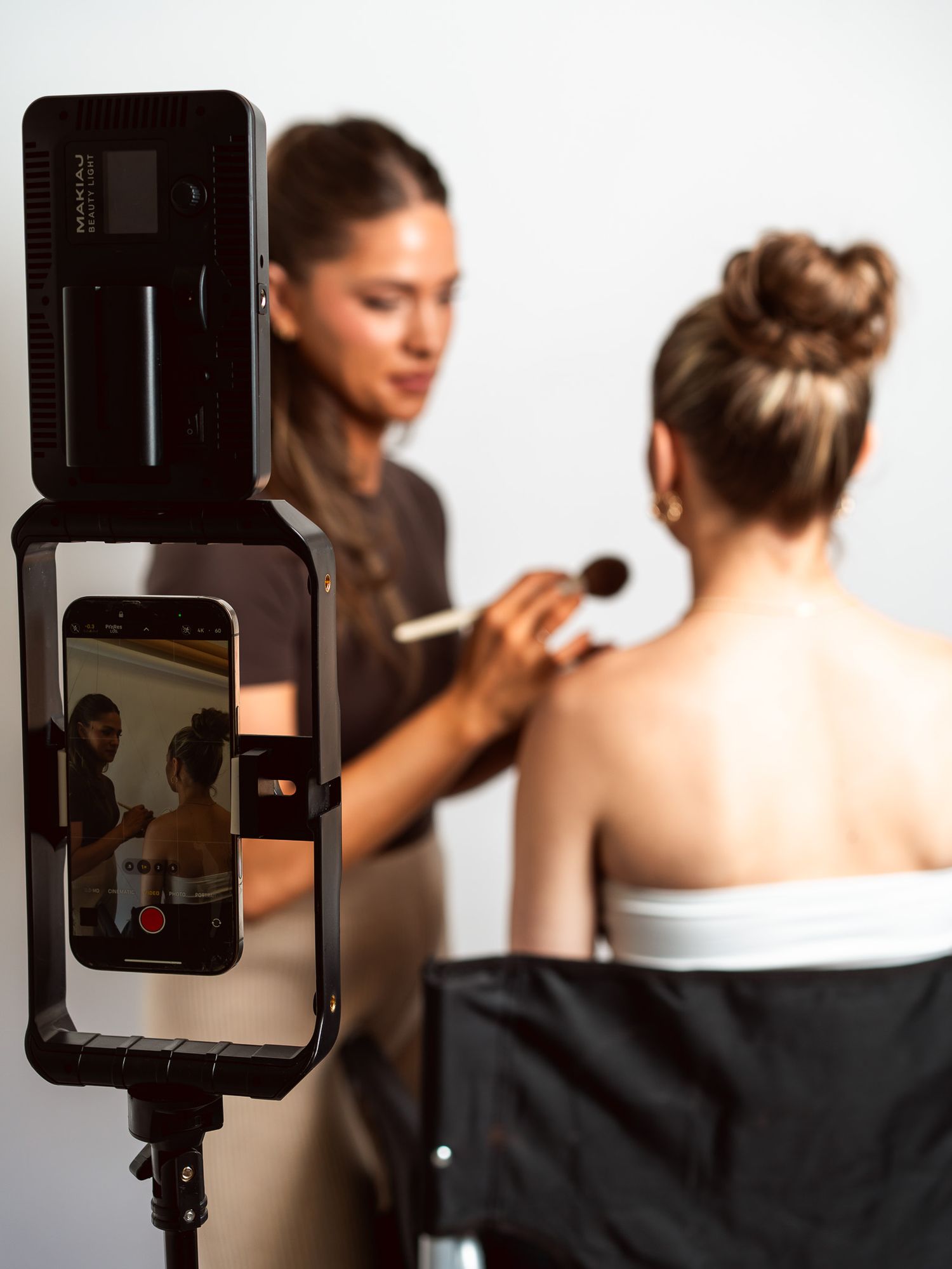Behind-the-scenes view of makeup application being recorded on a smartphone mounted to professional lighting equipment.