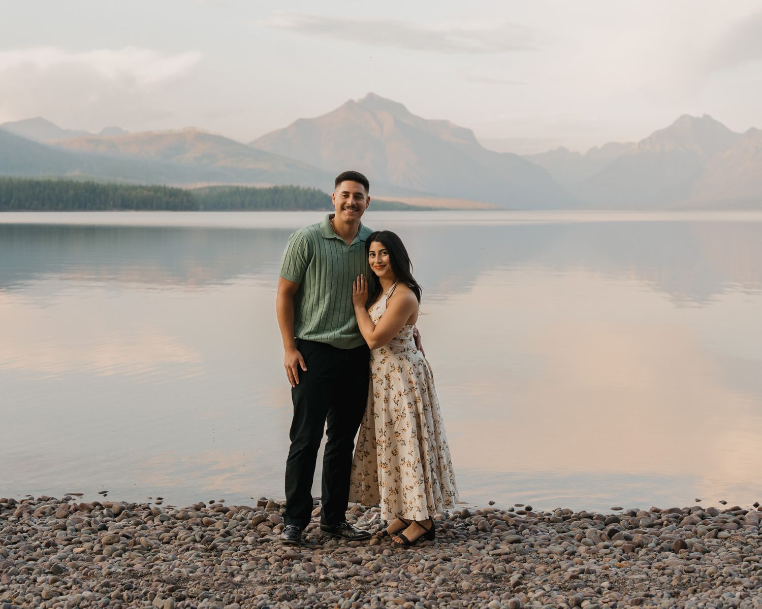 Couple smiling by a mountain lake at sunset, woman in floral dress, man in green shirt, rocky shoreline.