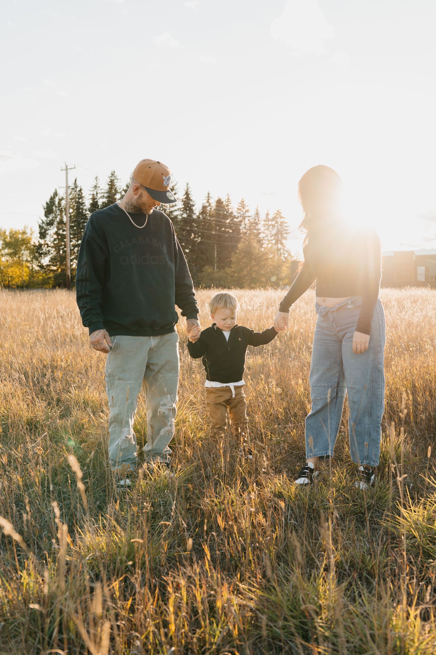 A family of three walks through a golden sunlit field, parents holding their toddler's hands at sunset.