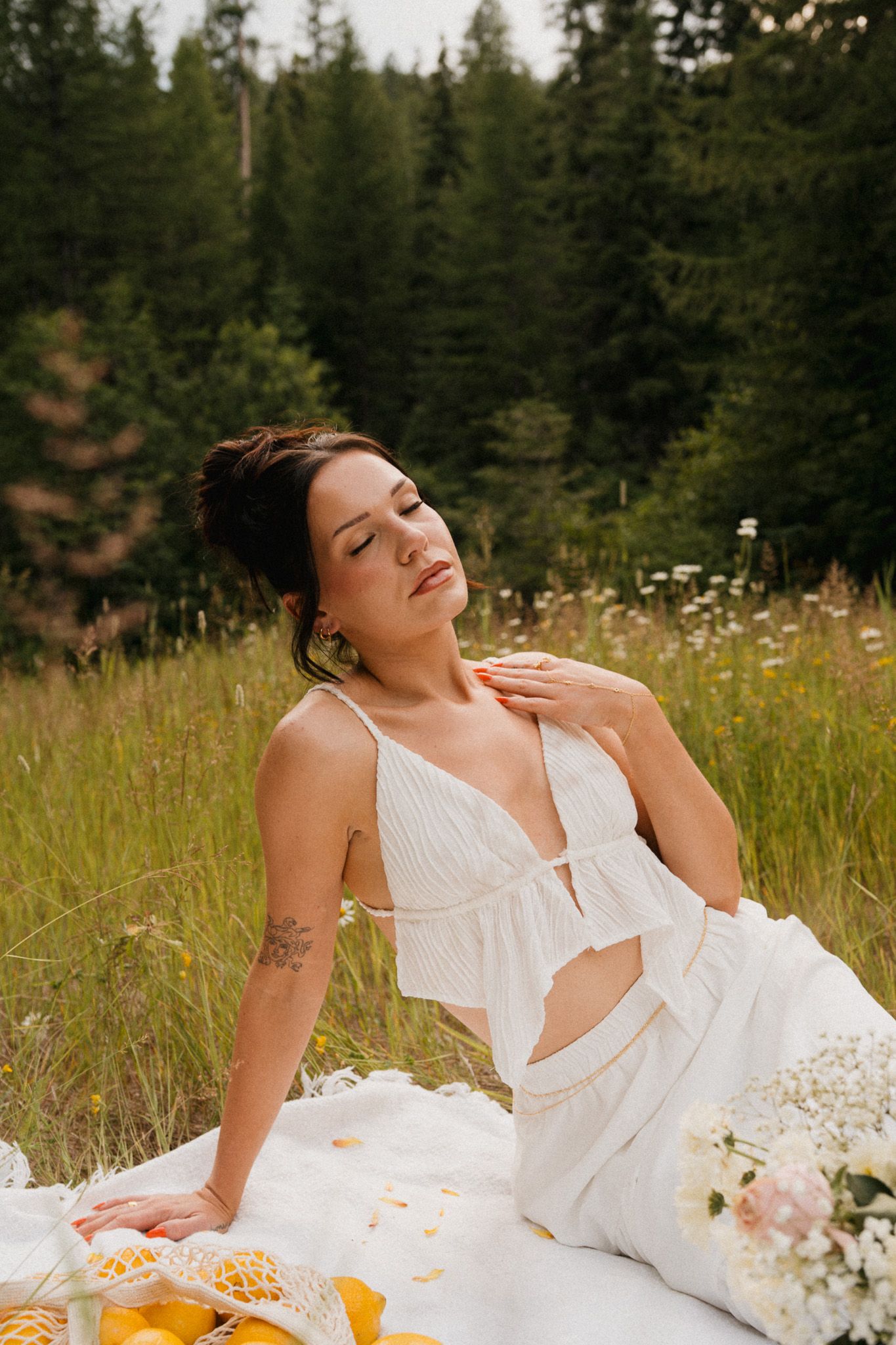 Woman in white crop top and skirt sitting on blanket in meadow with wildflowers and evergreen forest backdrop.