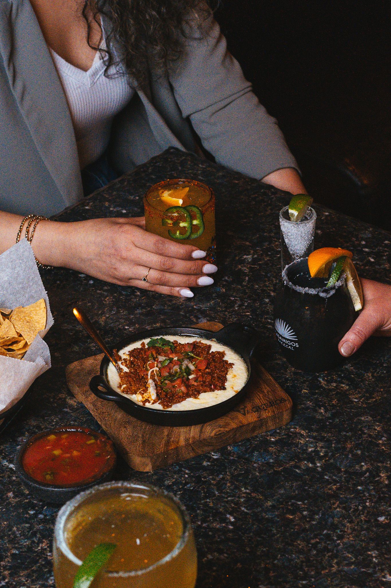 Mexican restaurant table spread with cocktails, sauces, and a meat dish on a wooden board against dark granite counter.