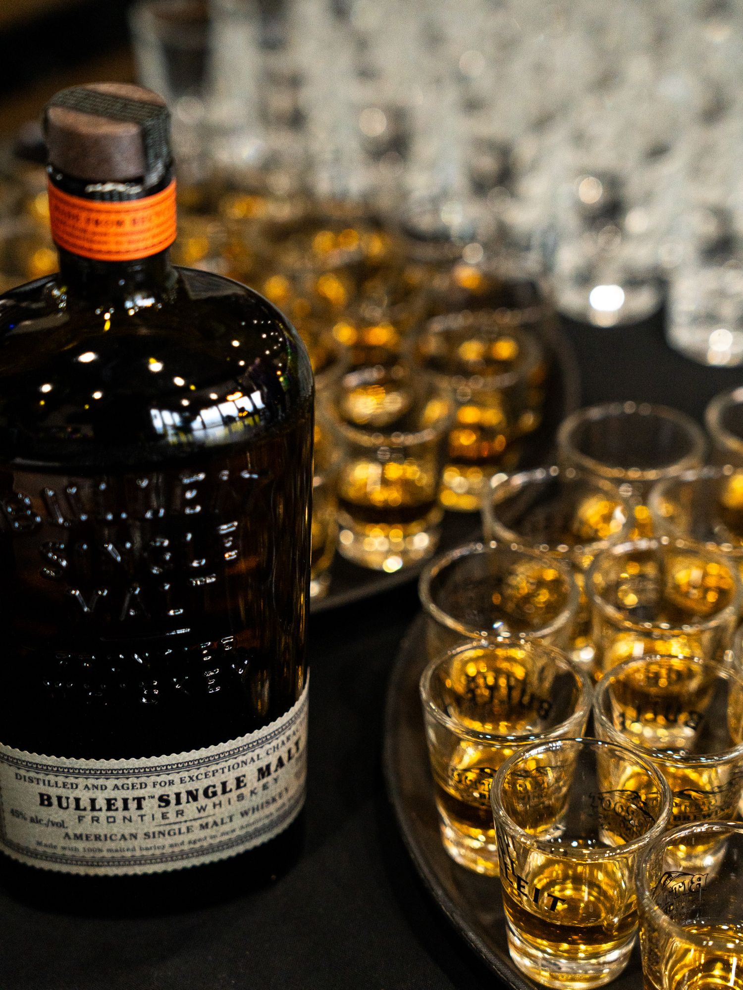 A bottle of Bulleit Single Malt whiskey with golden glasses arranged on a dark surface.