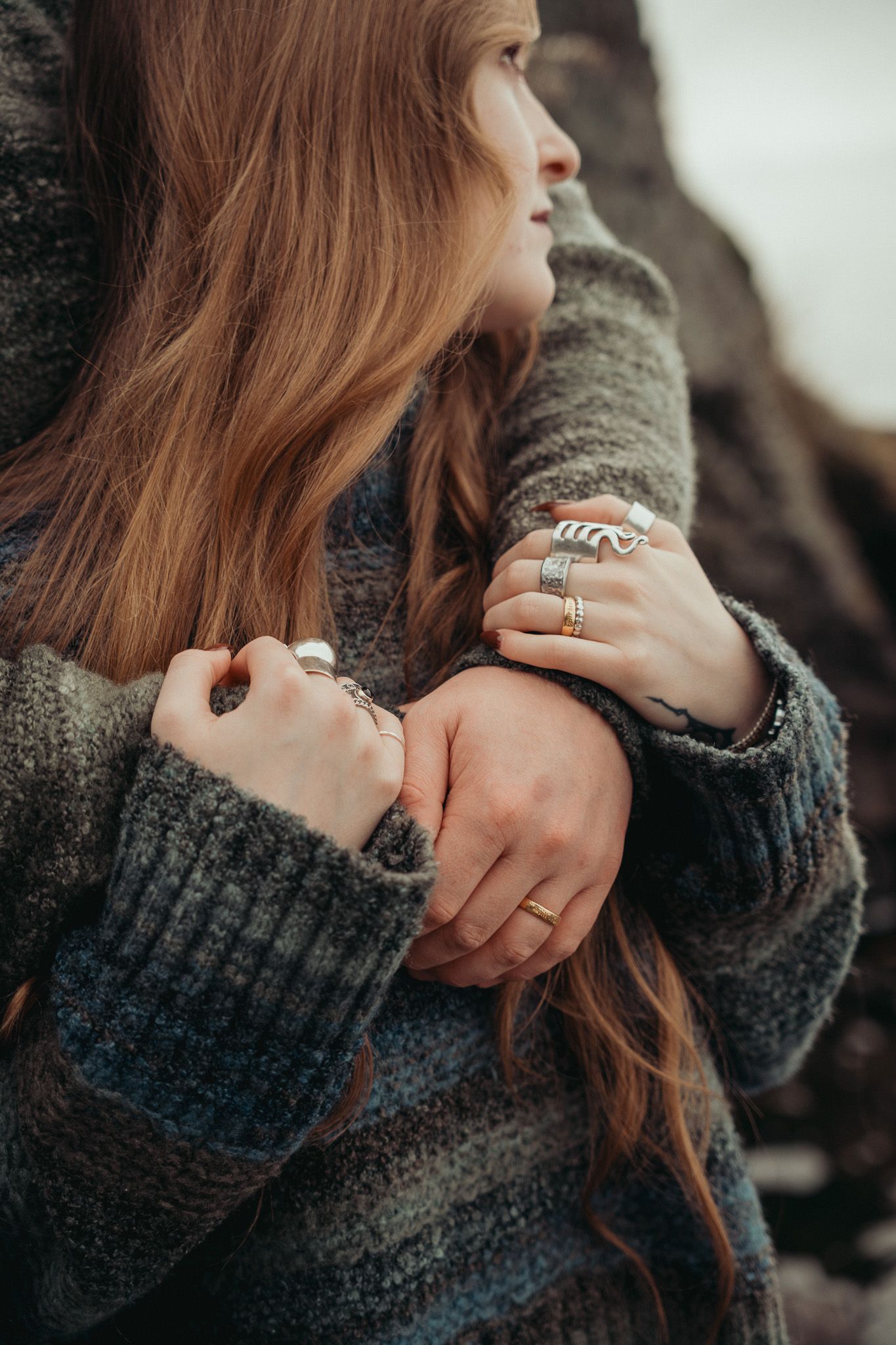 Woman in gray knit sweater with long auburn hair wearing stacked silver and gold rings, arms crossed, looking away outdoors.