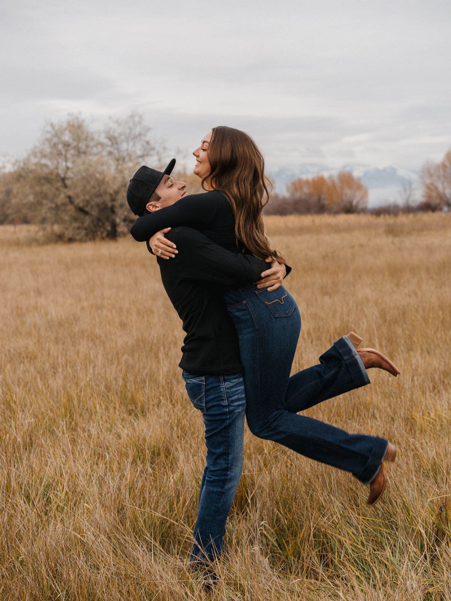 A joyful couple embraces in an autumn field, man lifting woman as she laughs, both wearing jeans and casual tops.
