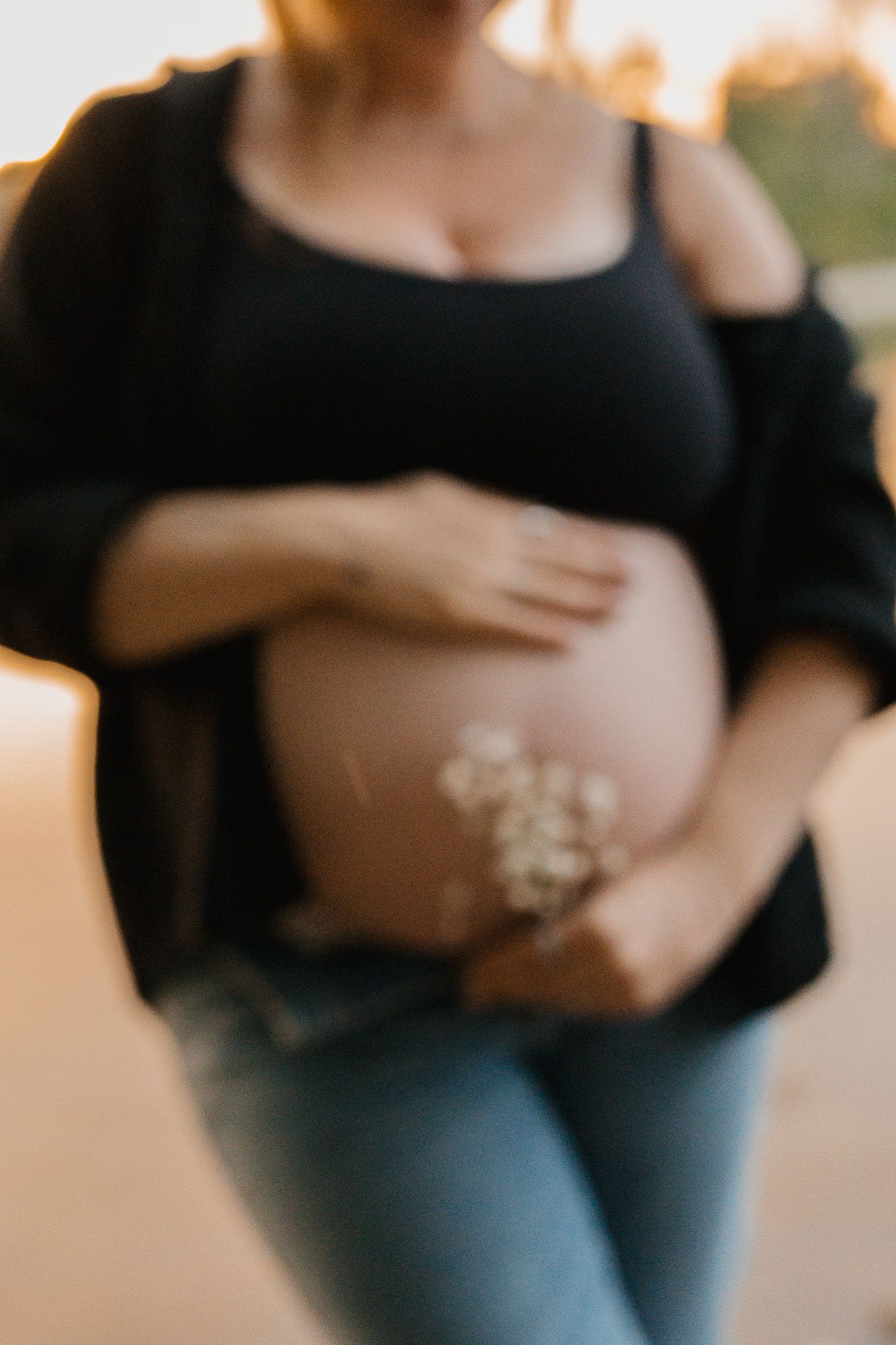Pregnant woman in black top and jeans cradling baby bump with small white flowers outdoors.