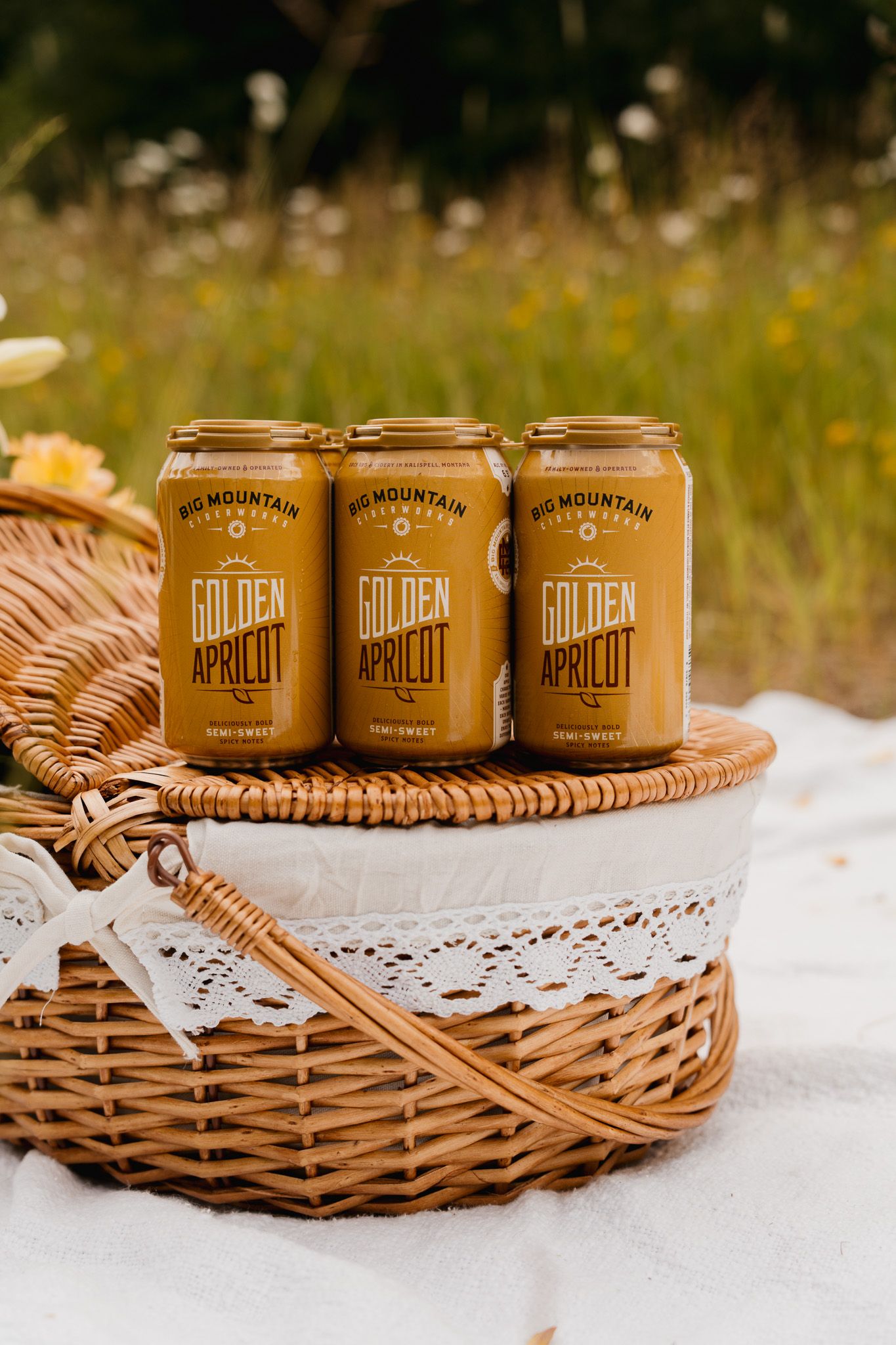 Three Golden Apricot canned drinks arranged on a wicker picnic basket with lace trim on a sunny summer day.