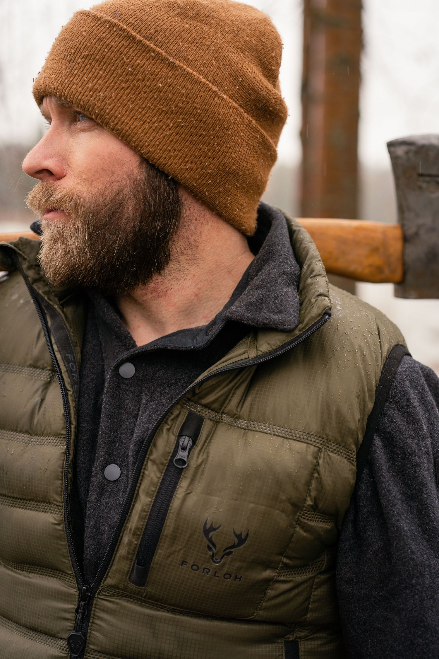 Person wearing brown beanie and olive puffer vest in outdoor winter setting.
