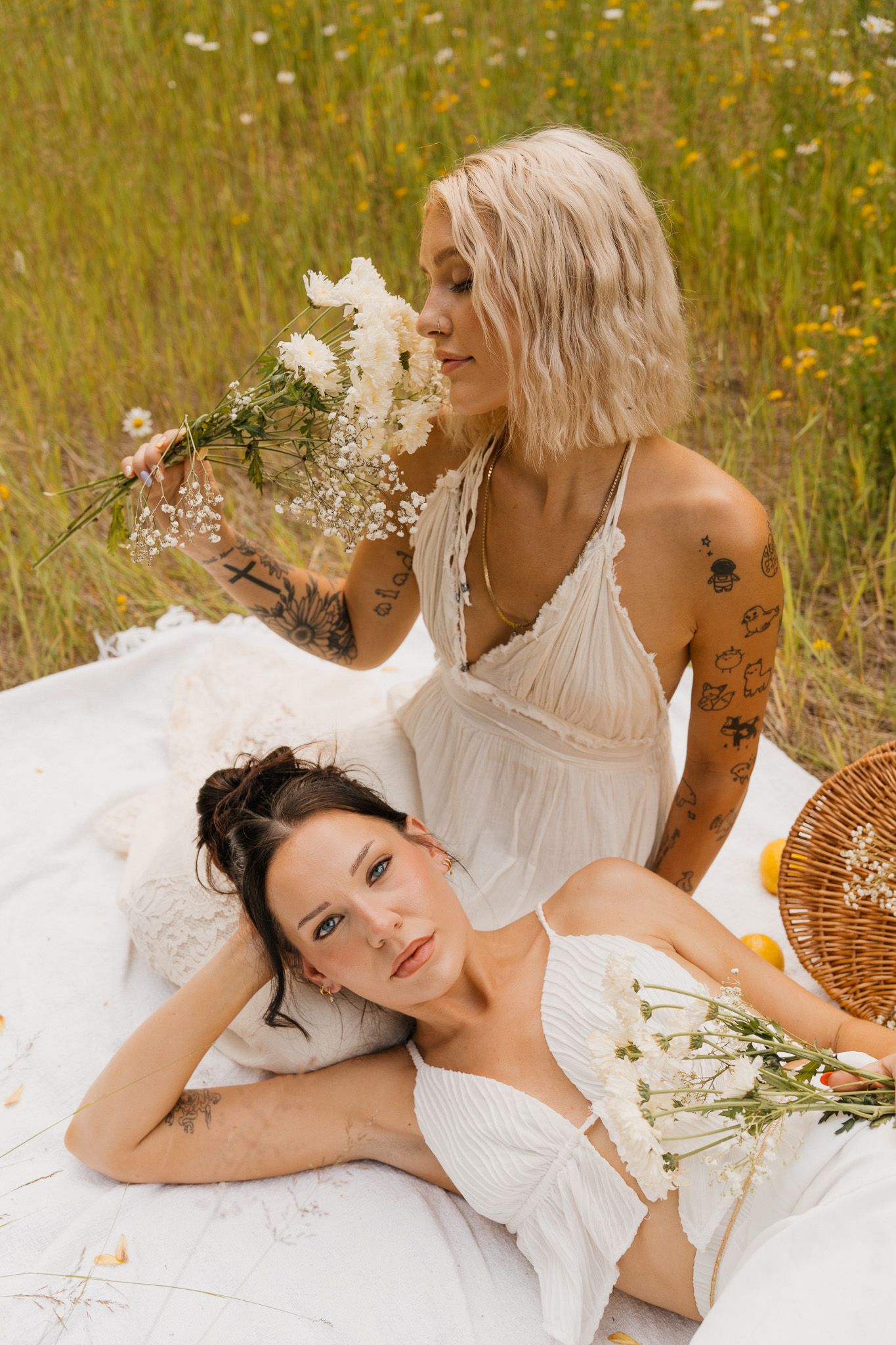 Two tattooed women in white dresses enjoy a summer picnic in a wildflower field with a wicker basket and white blooms.