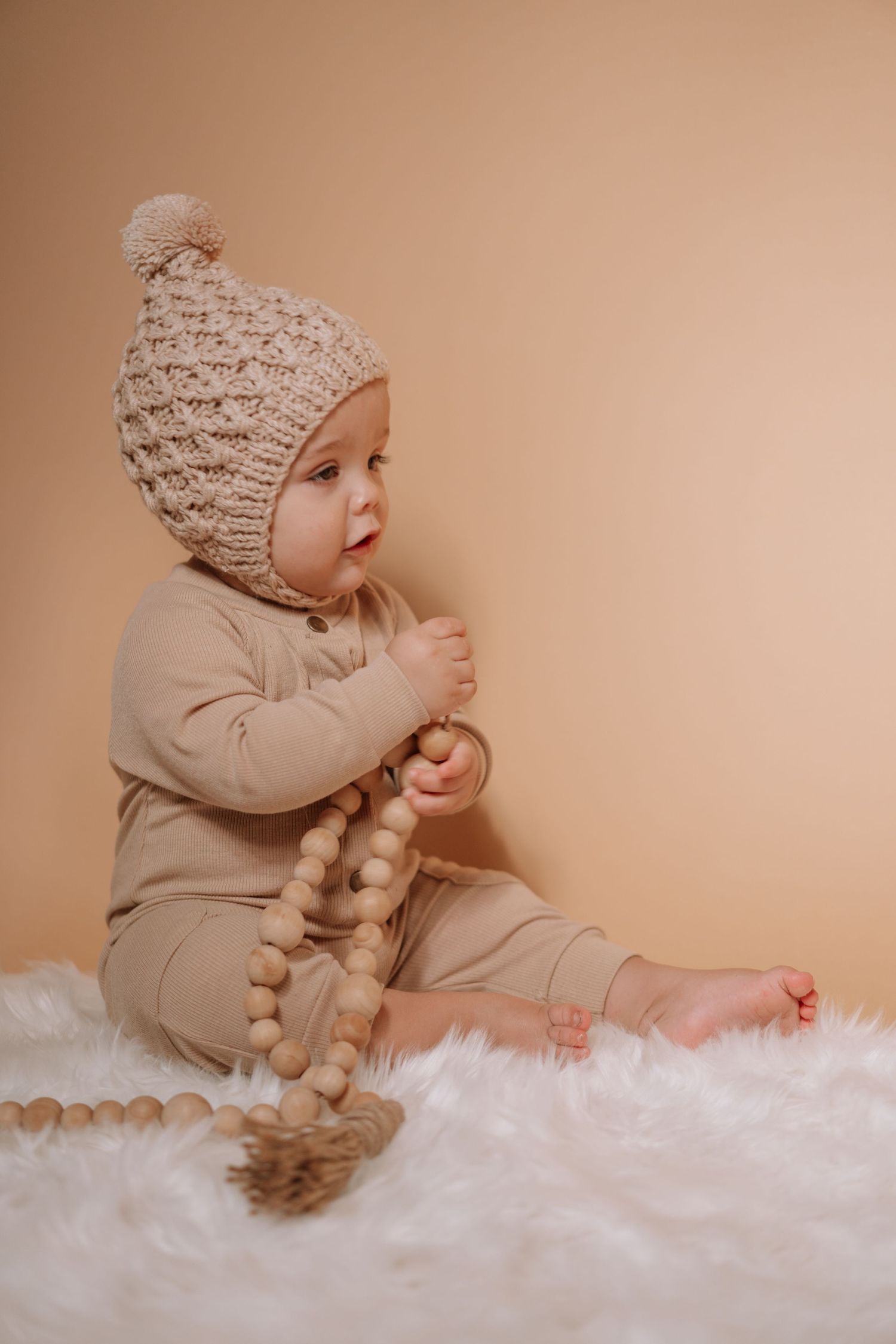 Baby in beige outfit and knit hat sitting on white fur rug with wooden bead garland.