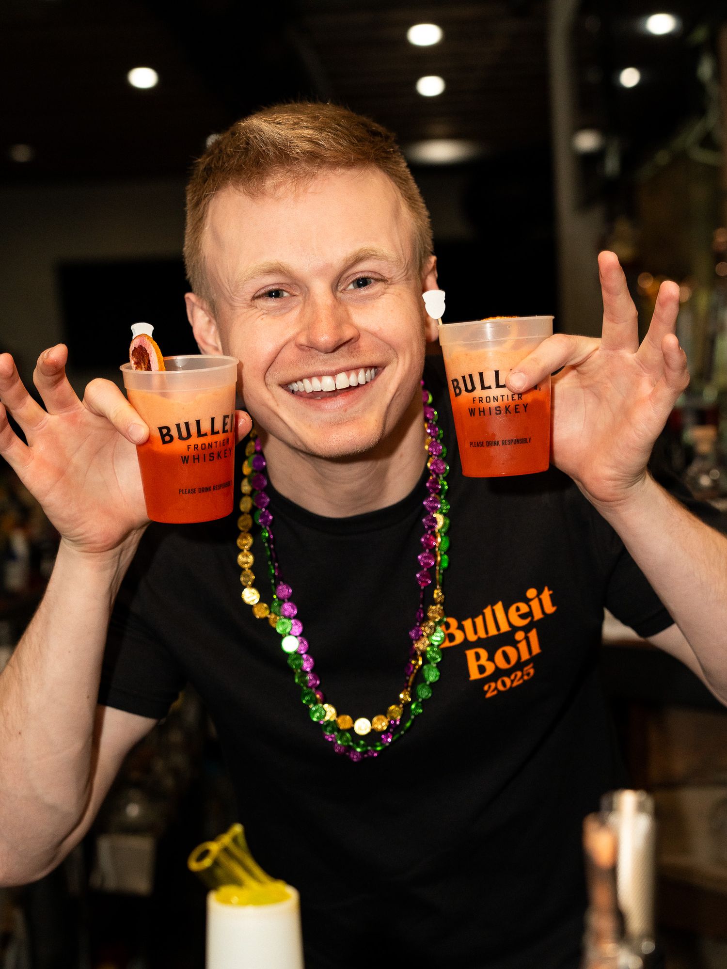 Smiling server in black Bulleit shirt holding cocktails and wearing colorful Mardi Gras beads.