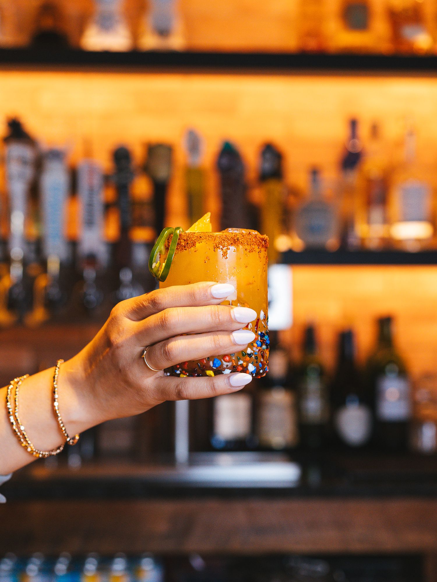 Cocktail being served at a bar with warm ambient lighting and bottles in background.