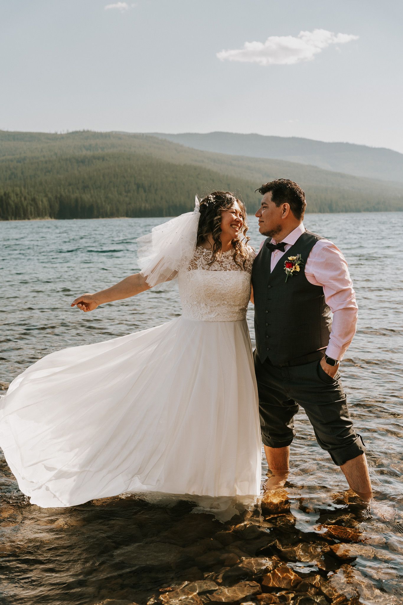 Bride and groom standing in a mountain lake, bride in white tulle gown, groom in vest and pink shirt, smiling at each other.