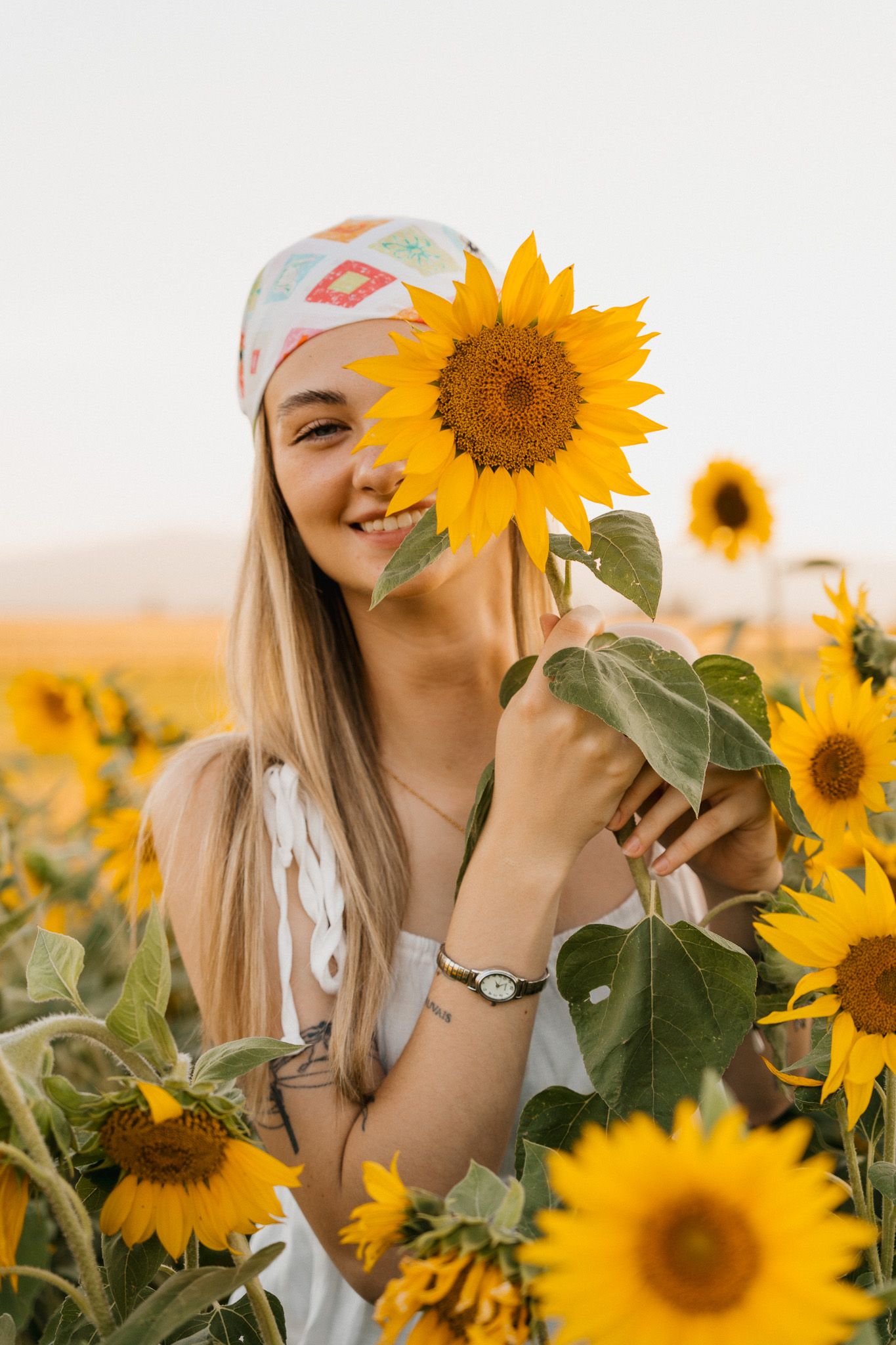 Smiling young woman holding a sunflower in a vibrant sunflower field, wearing a white dress and patterned headband.