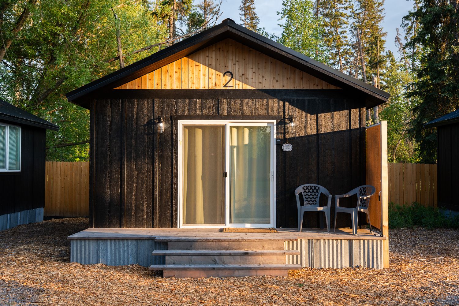 Modern wood cabin exterior with black roof and large windows nestled among tall pine trees in a forest setting.