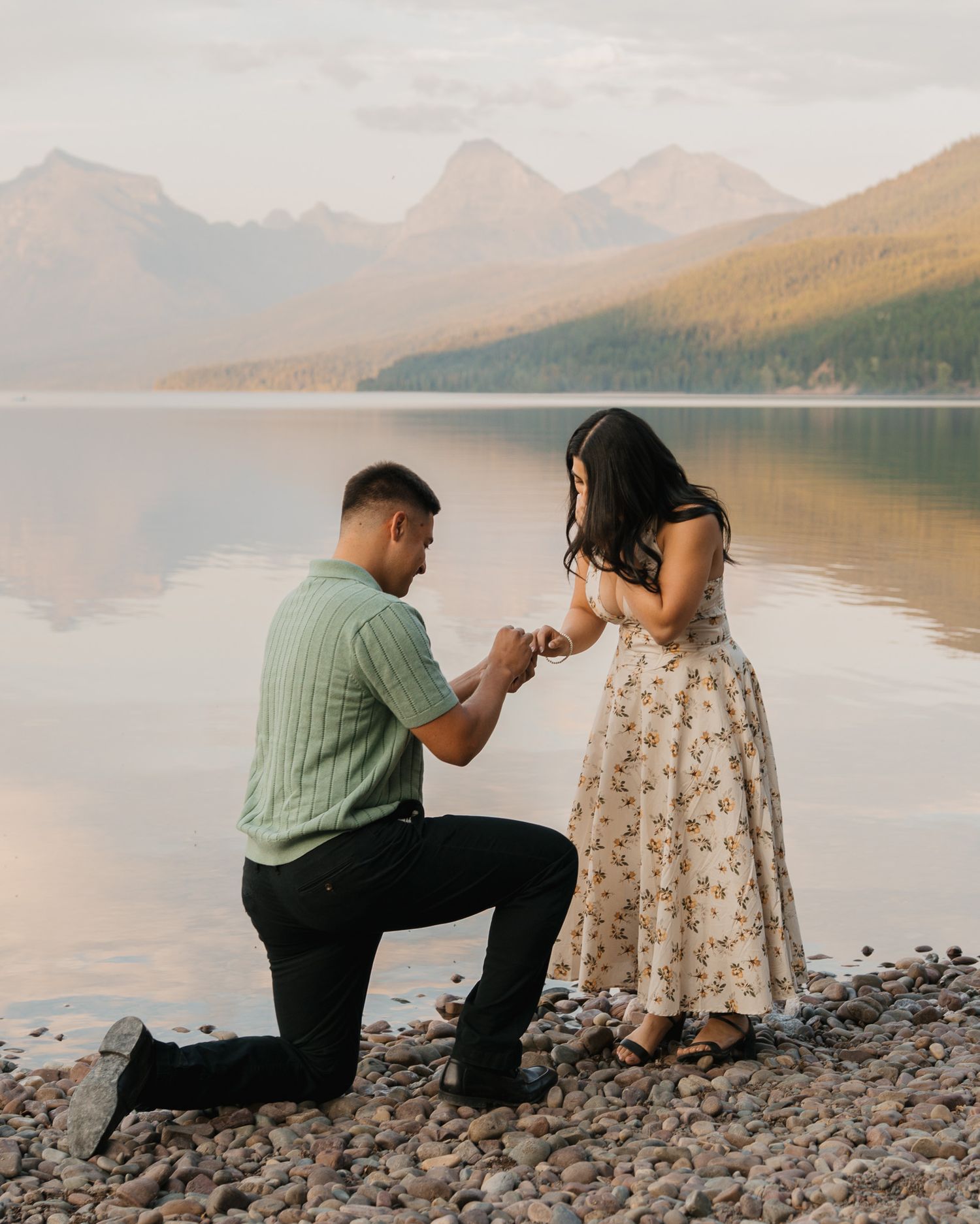 Man proposes on one knee to woman by a serene mountain lake at sunset on a rocky shore.