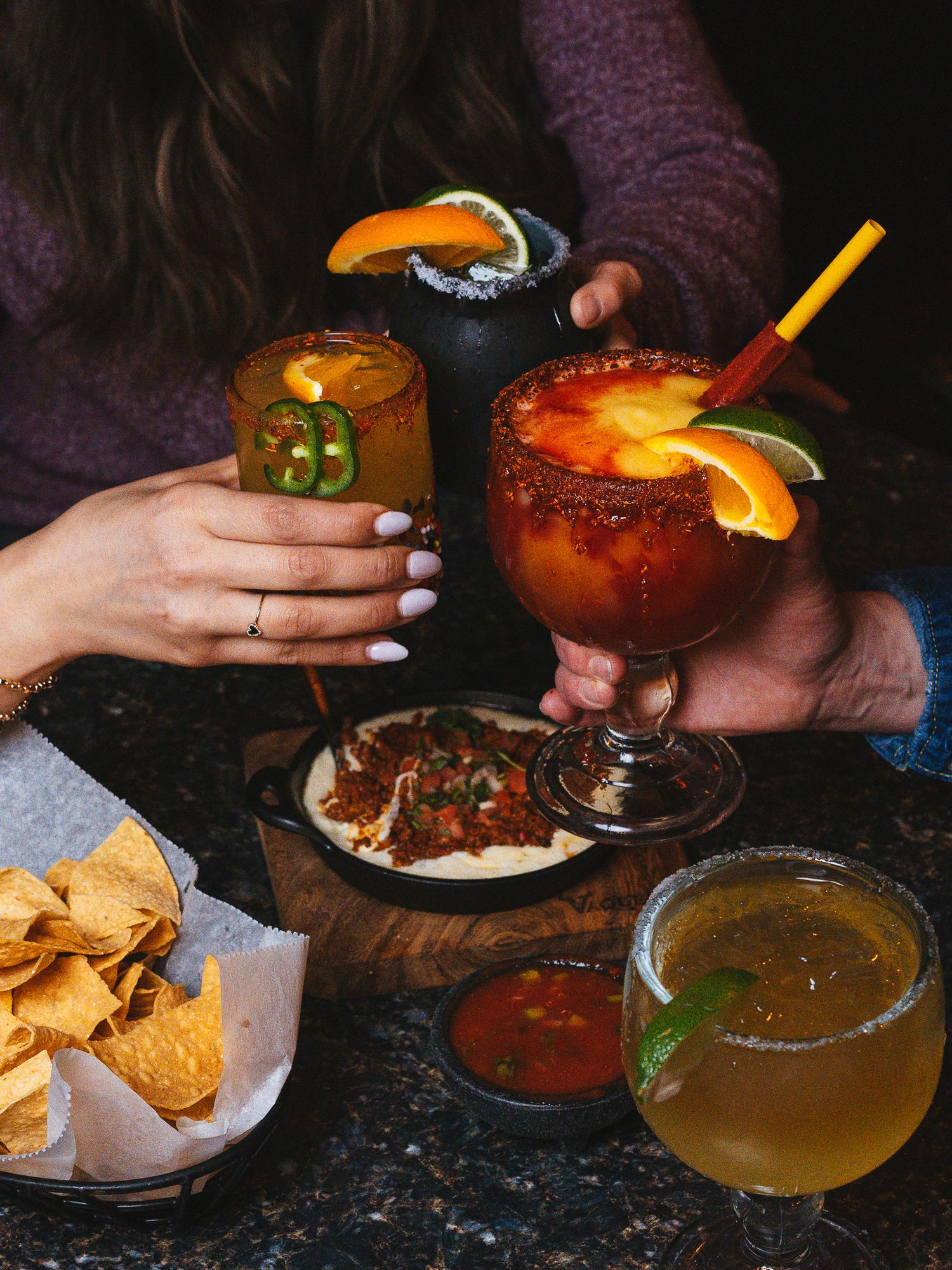 Colorful cocktails and Mexican food spread with tortilla chips, sauces and garnishes on dark table.