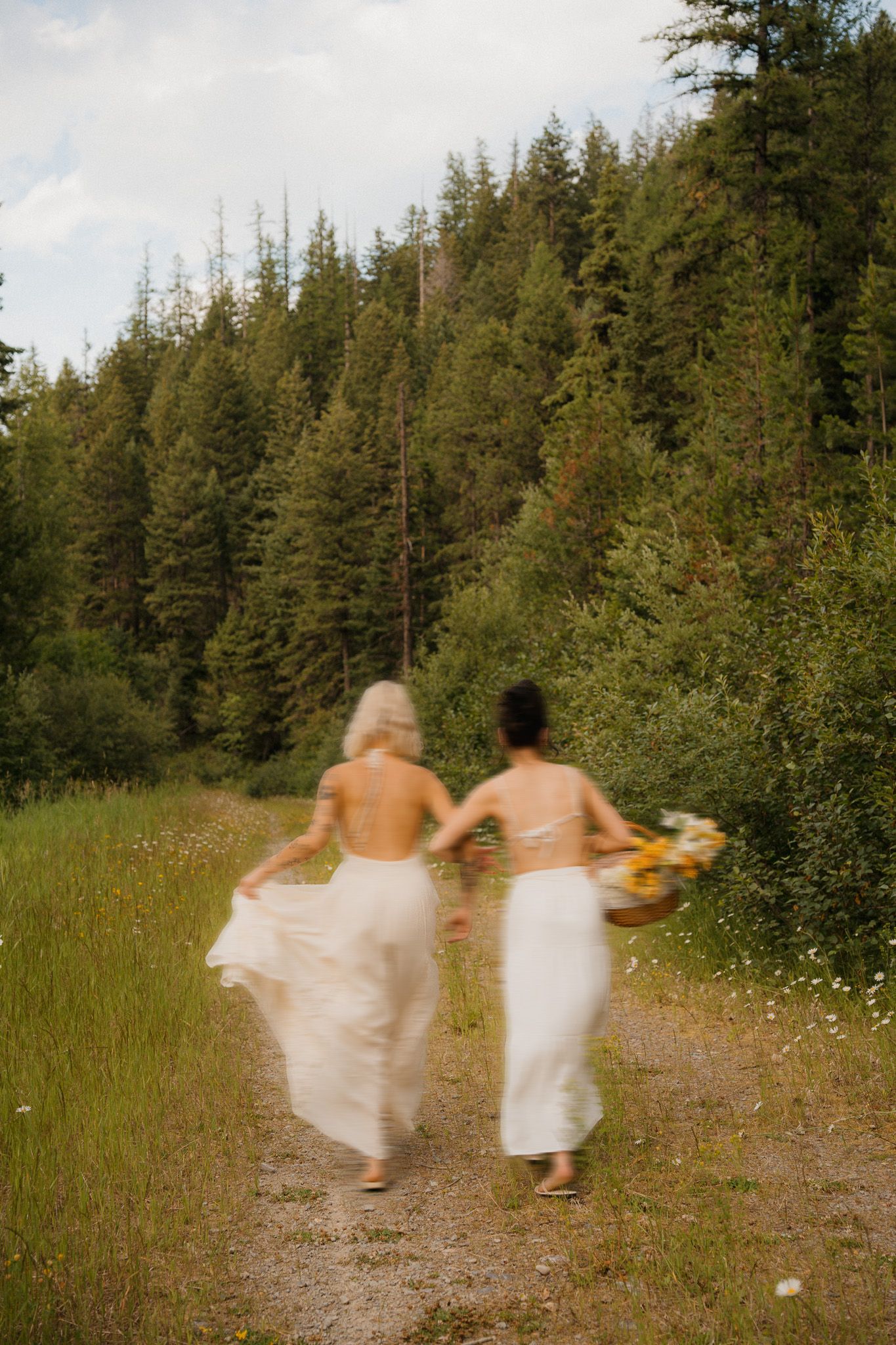 Two brides in white dresses hold hands running down a forest path, one carrying a wildflower bouquet.