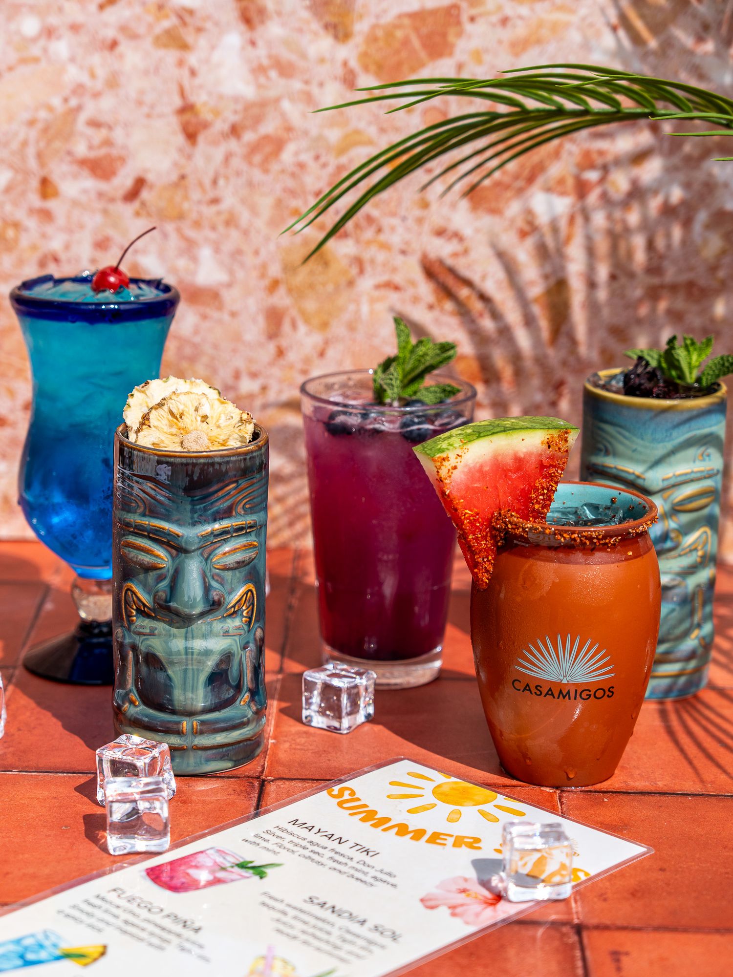Tropical tiki cocktails in decorative glasses with fruit garnishes and menu on coral-colored table.