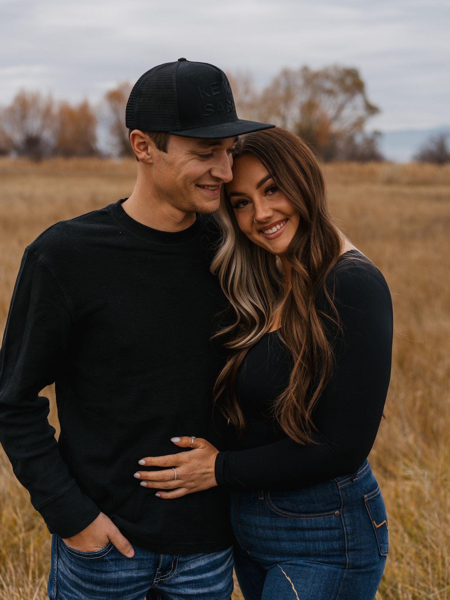 A couple in matching black outfits embraces warmly in an autumn field, smiling together for a romantic outdoor photo.