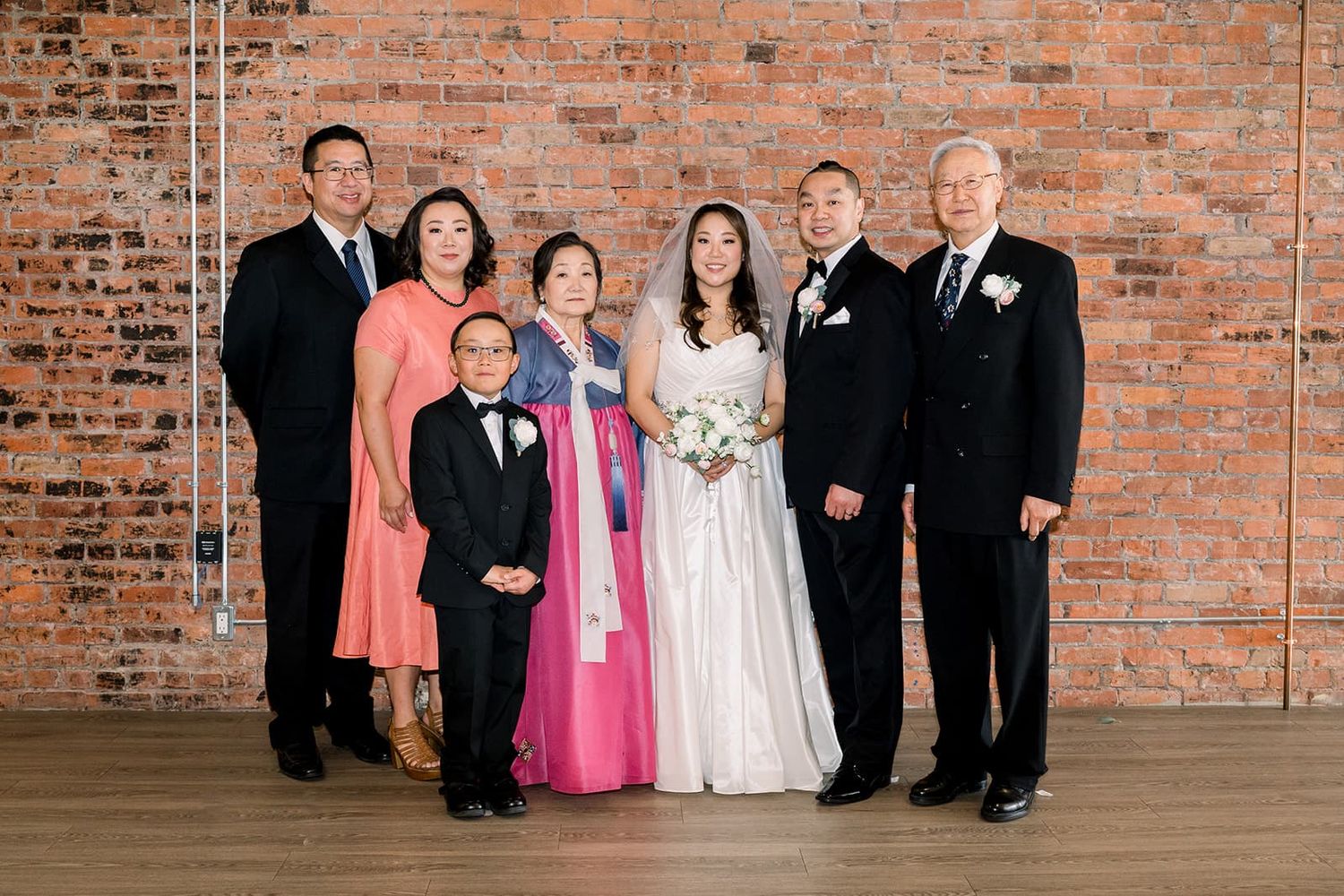 Wedding family portrait of seven people posing together in front of a rustic brick wall.