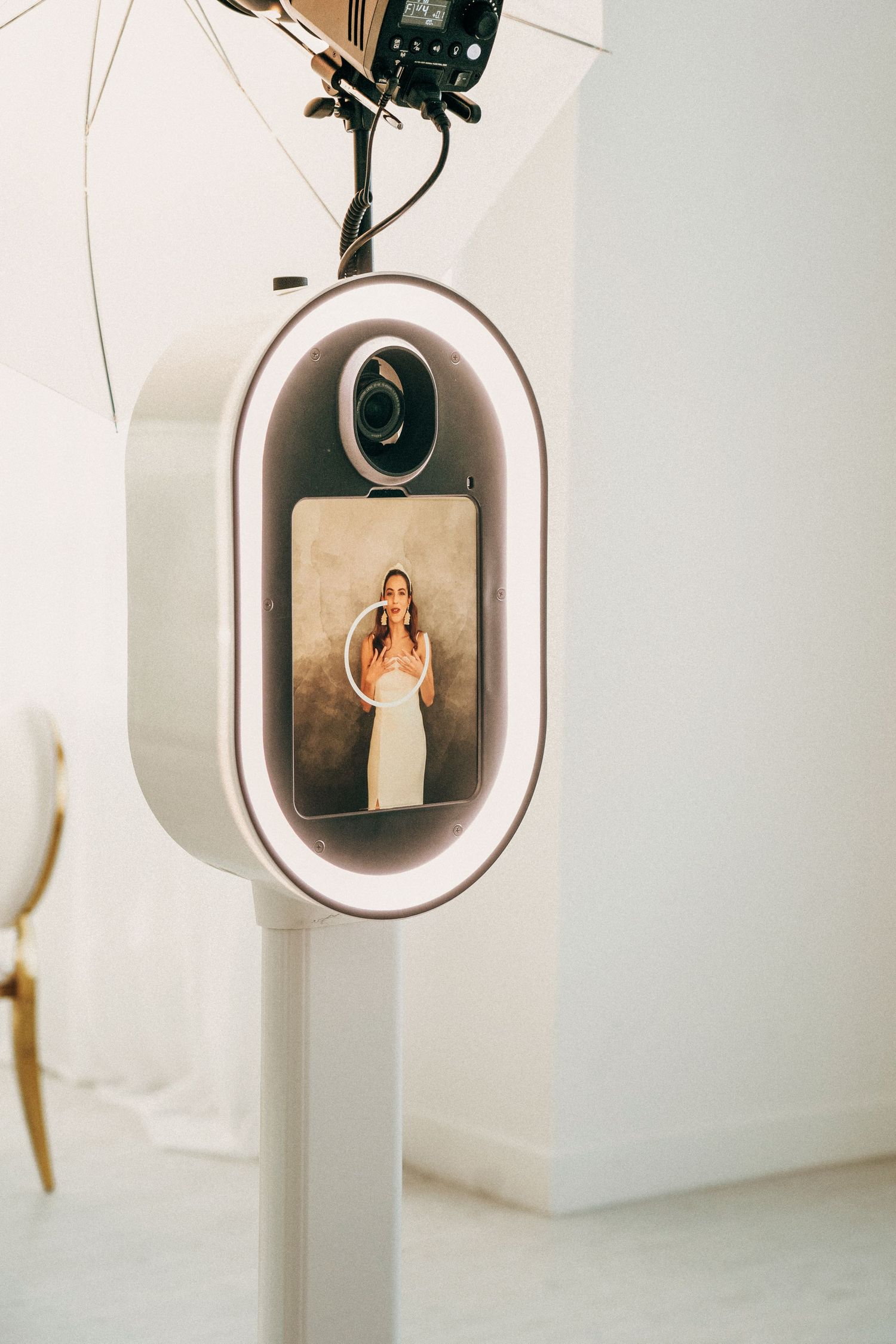 A modern photo booth with an oval-shaped LED frame mounted on a white stand displays a photo on its screen.