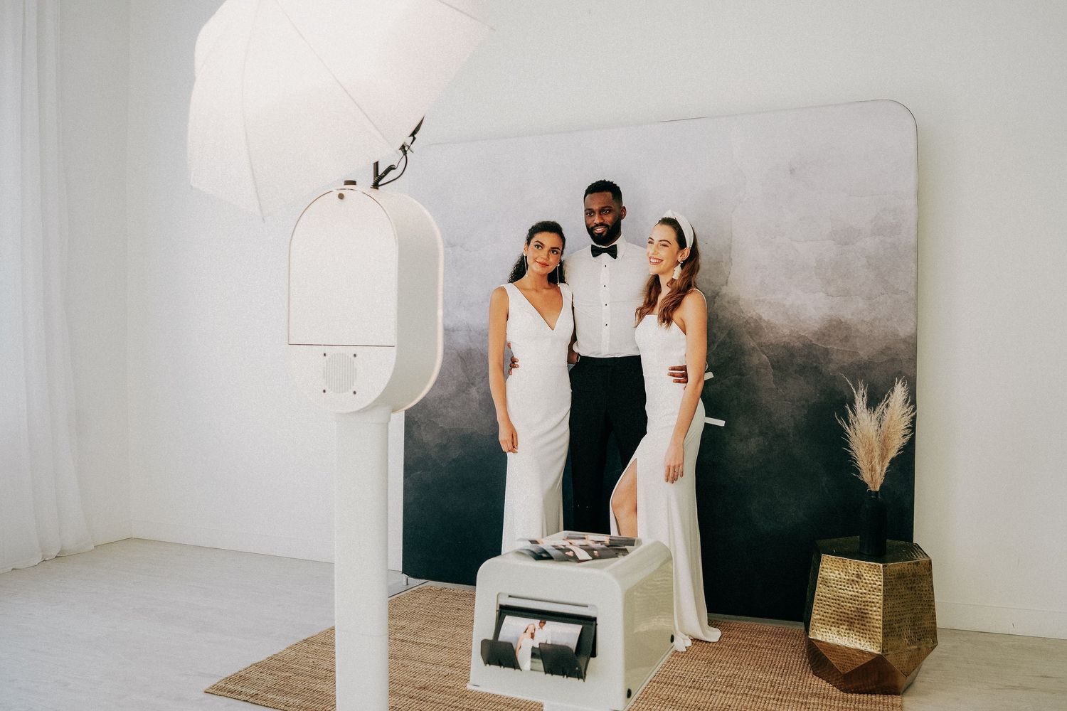 Three people in elegant white outfits pose together against a dark wall with minimalist decor in a bright studio setting.