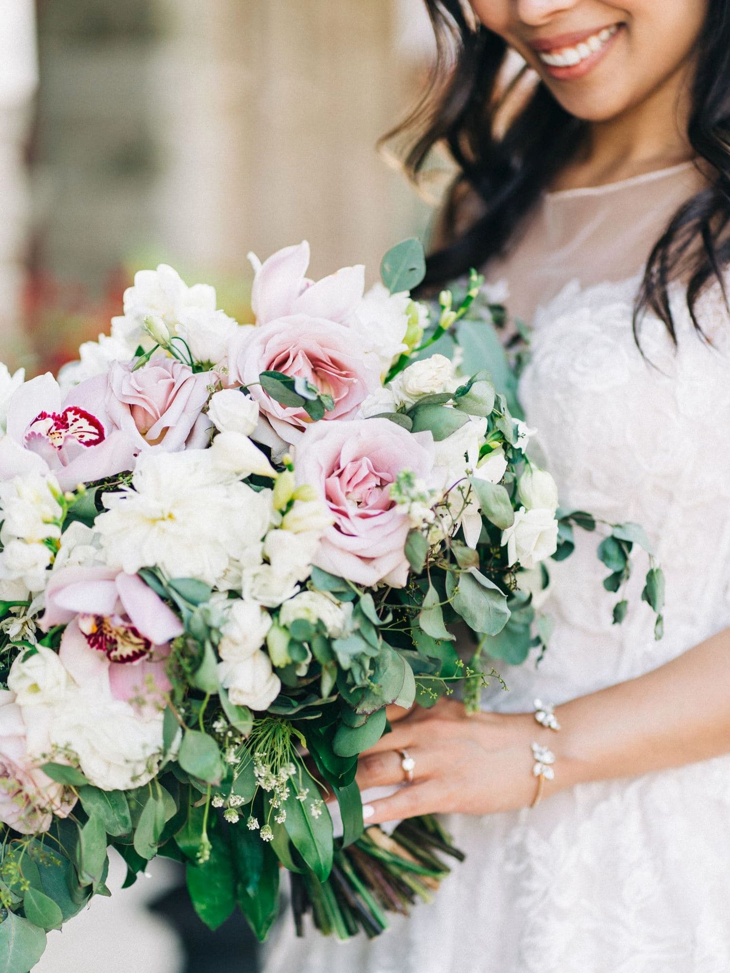 Close-up of a bride holding a lush bouquet of pink roses, orchids, and greenery in a lace wedding dress.