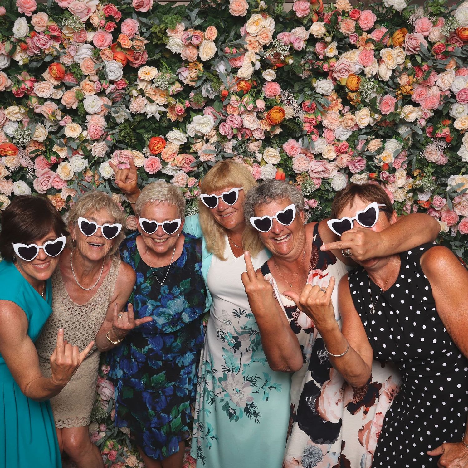 Six women wearing heart-shaped sunglasses pose playfully in front of a pink floral wall.