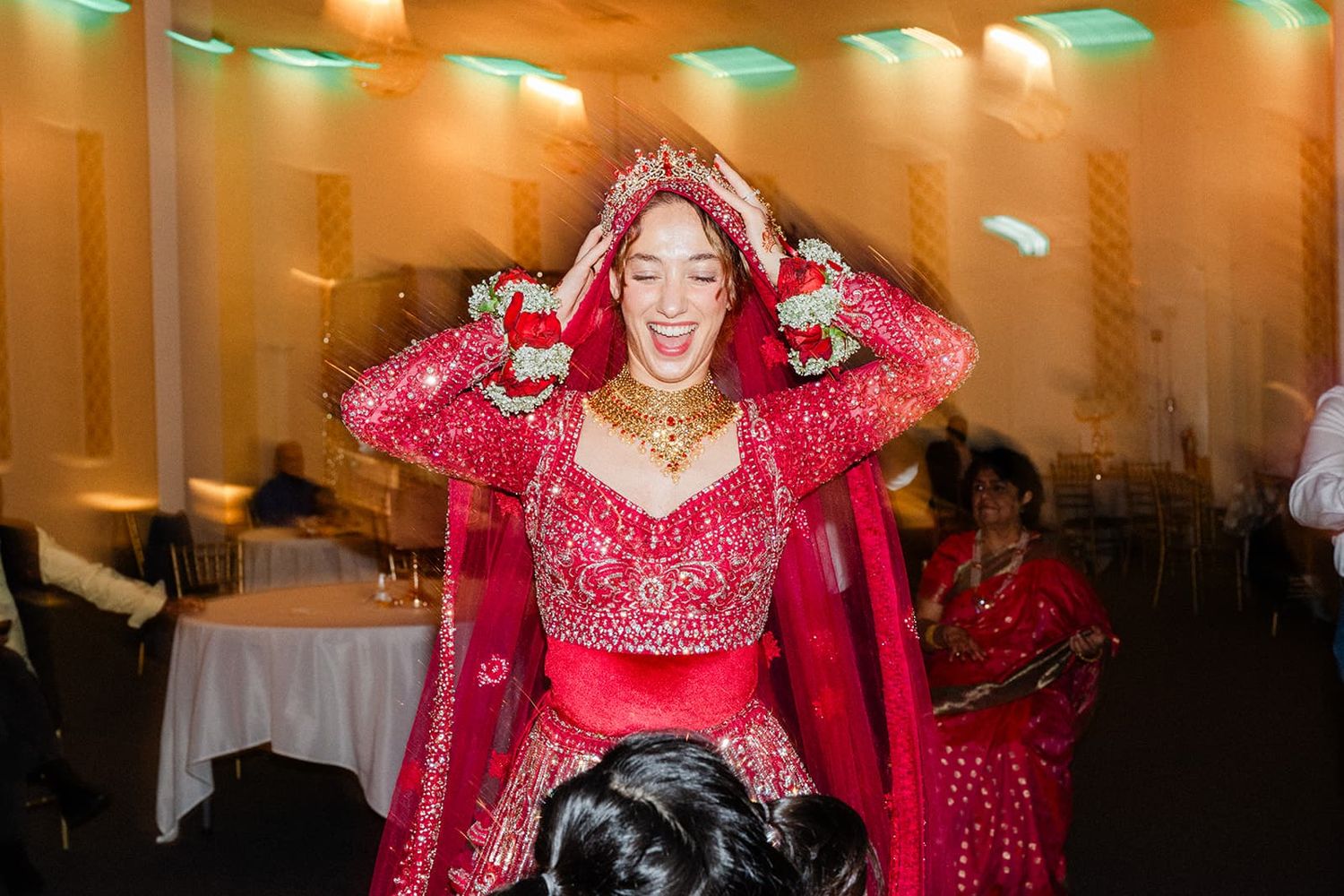 Joyful bride in red embroidered lehenga and floral accessories laughs during traditional Indian wedding celebration.