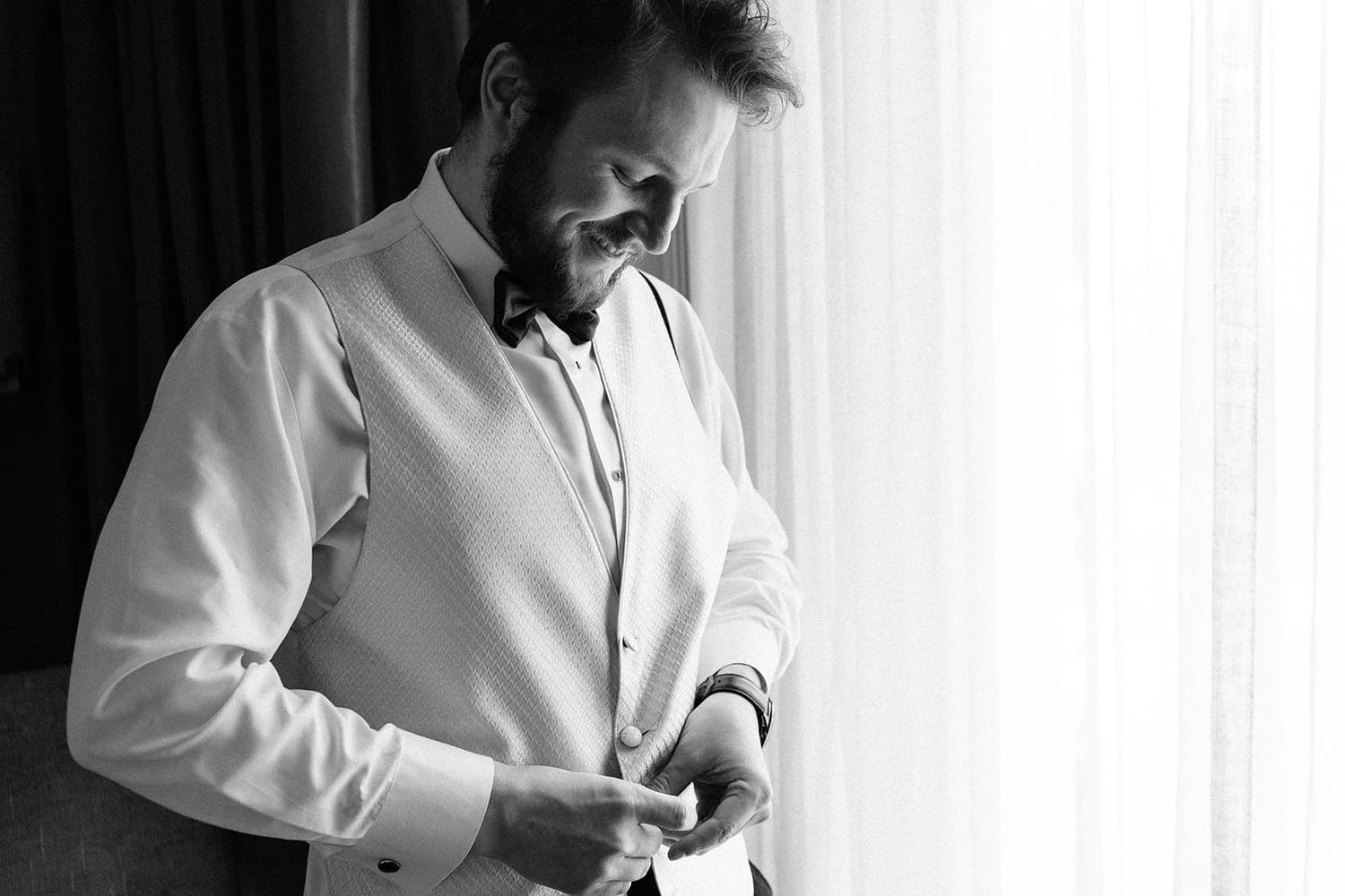 Groom in white dress shirt buttoning his cuff while getting ready for his wedding day.