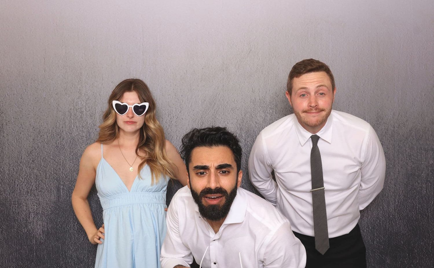 Three friends pose in a photo booth, with a woman in a blue dress wearing cat-eye sunglasses.