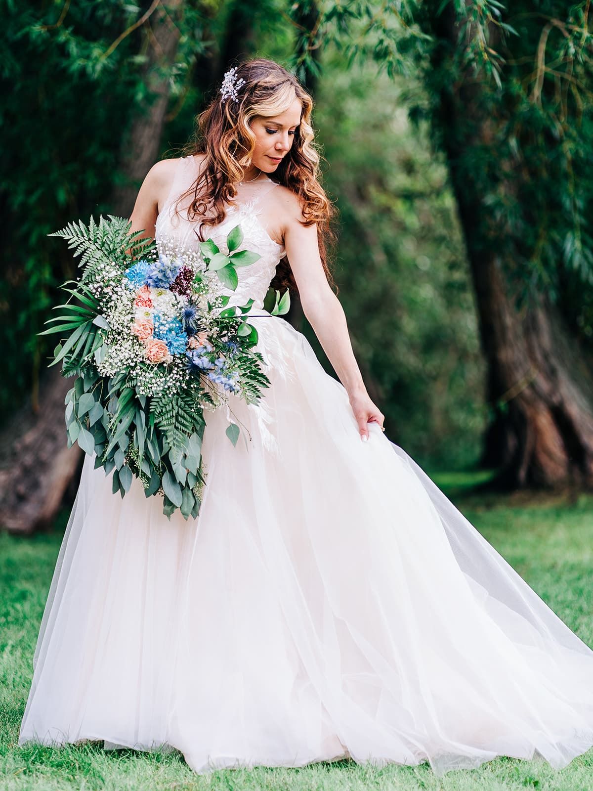 Bride in a blush tulle ball gown holding a lush bouquet of blue and green flowers, posing outdoors among tall trees.
